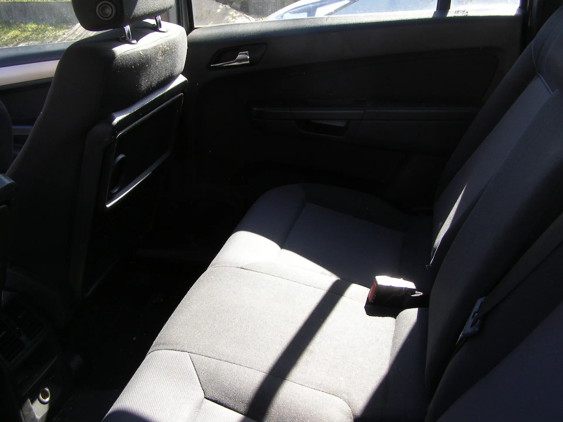 Wnętrze samochodu z widokiem na tylne siedzenie. Czarne siedzenia z poduszką podłokietnika po lewej stronie, uchwyt na drzwiach, pas bezpieczeństwa z czerwonym elementem zapięcia. Przez okno widoczne są zewnętrzne elementy: trawa, fragment innego pojazdu i betonowa powierzchnia. Światło słoneczne rzuca cienie na siedzenie.