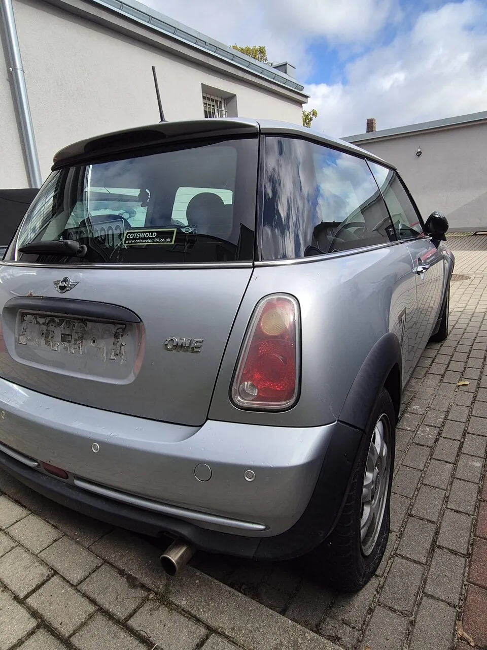Srebrny samochód marki Mini Cooper model One, zaparkowany tyłem na kostce brukowej. Na tylnej szybie znajduje się naklejka z napisem 'COTSWOLD www.cotswold@mail.co.uk'. W tle widoczny jest jasny budynek oraz niebo z chmurami.