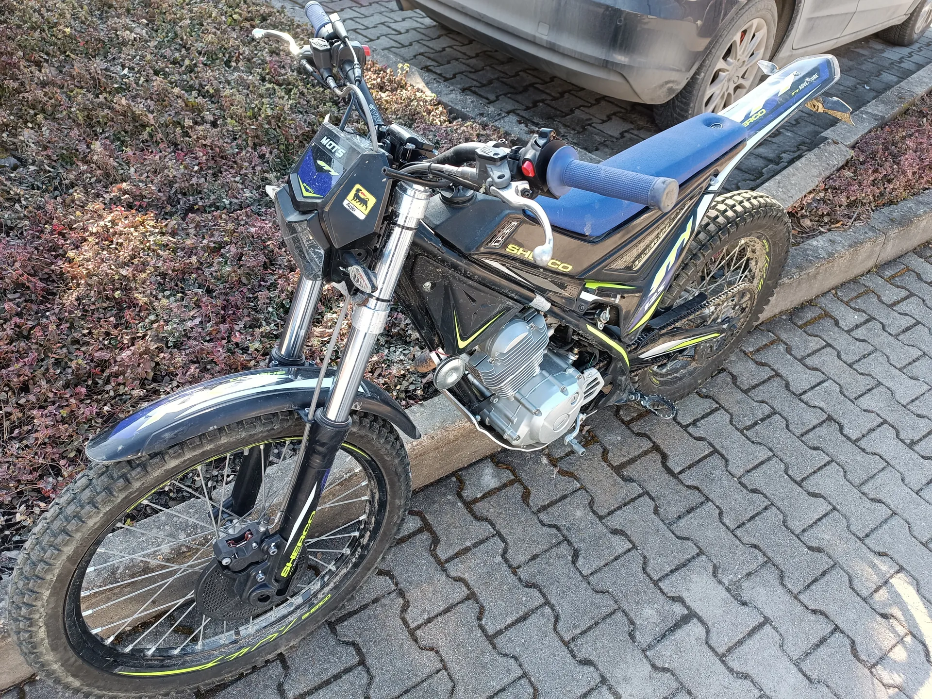 Motocykl marki Sherco w kolorze czarnym z niebieskimi i żółtymi akcentami, zaparkowany na brukowanej powierzchni. Widoczne są czarne opony z żółtymi detalami, niebieskie siedzenie, kierownica z niebieskimi uchwytami oraz tabliczka z napisem 'MOT5'. W tle krzewy z brązowymi liśćmi i fragment szarego samochodu. Motocykl stoi na chodniku z kamiennych płyt.