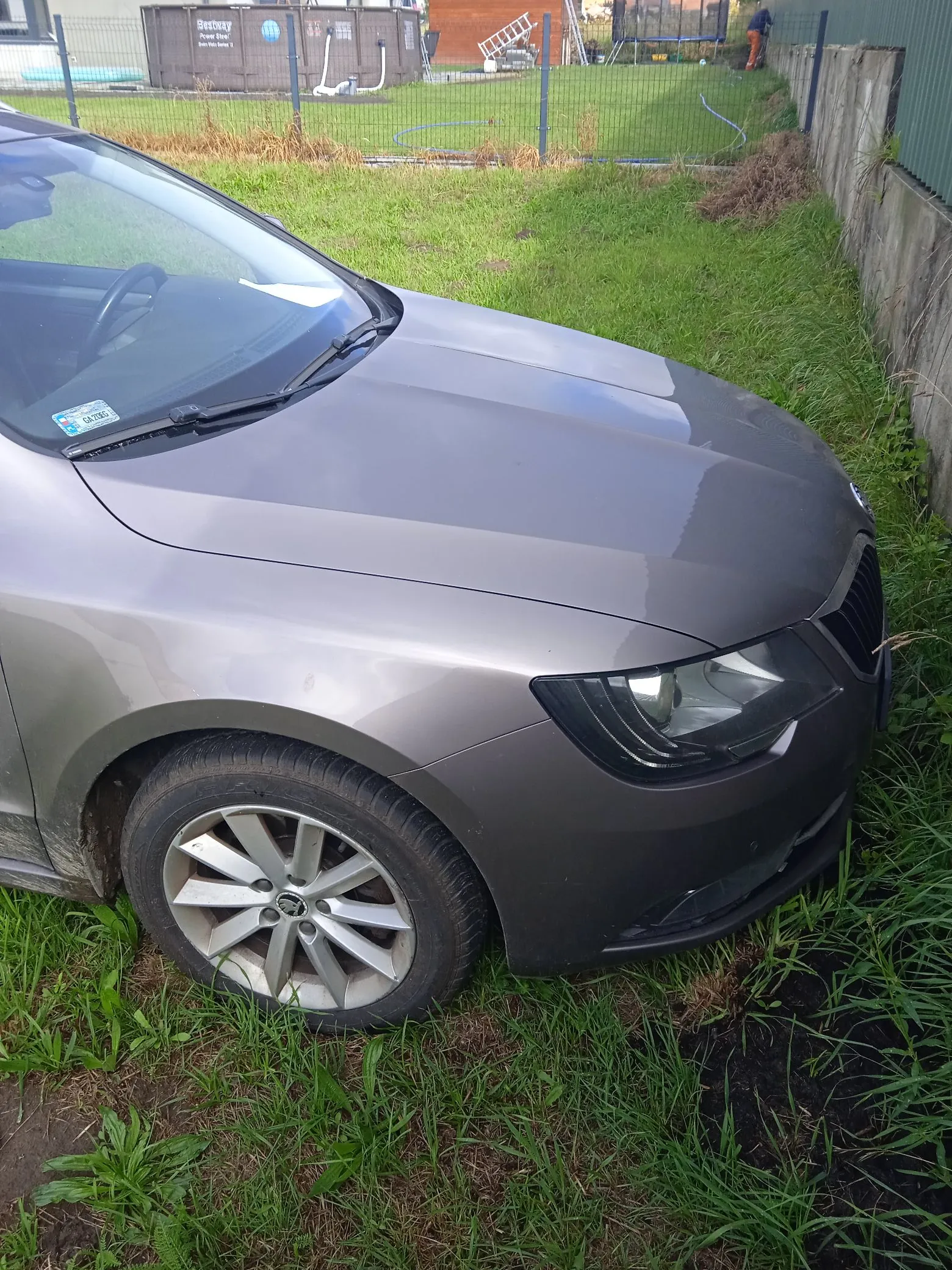 Zbliżenie na przód szarego samochodu osobowej marki Škoda, zaparkowanego na trawie. Widoczny jest fragment maski, przednia prawa lampa, przednie prawe koło z aluminianą felgą oraz fragment przedniej szyby z naklejką ubezpieczeniową. W tle znajduje się zielony trawnik, ogrodzenie z siatki oraz betonowy mur po prawej stronie.