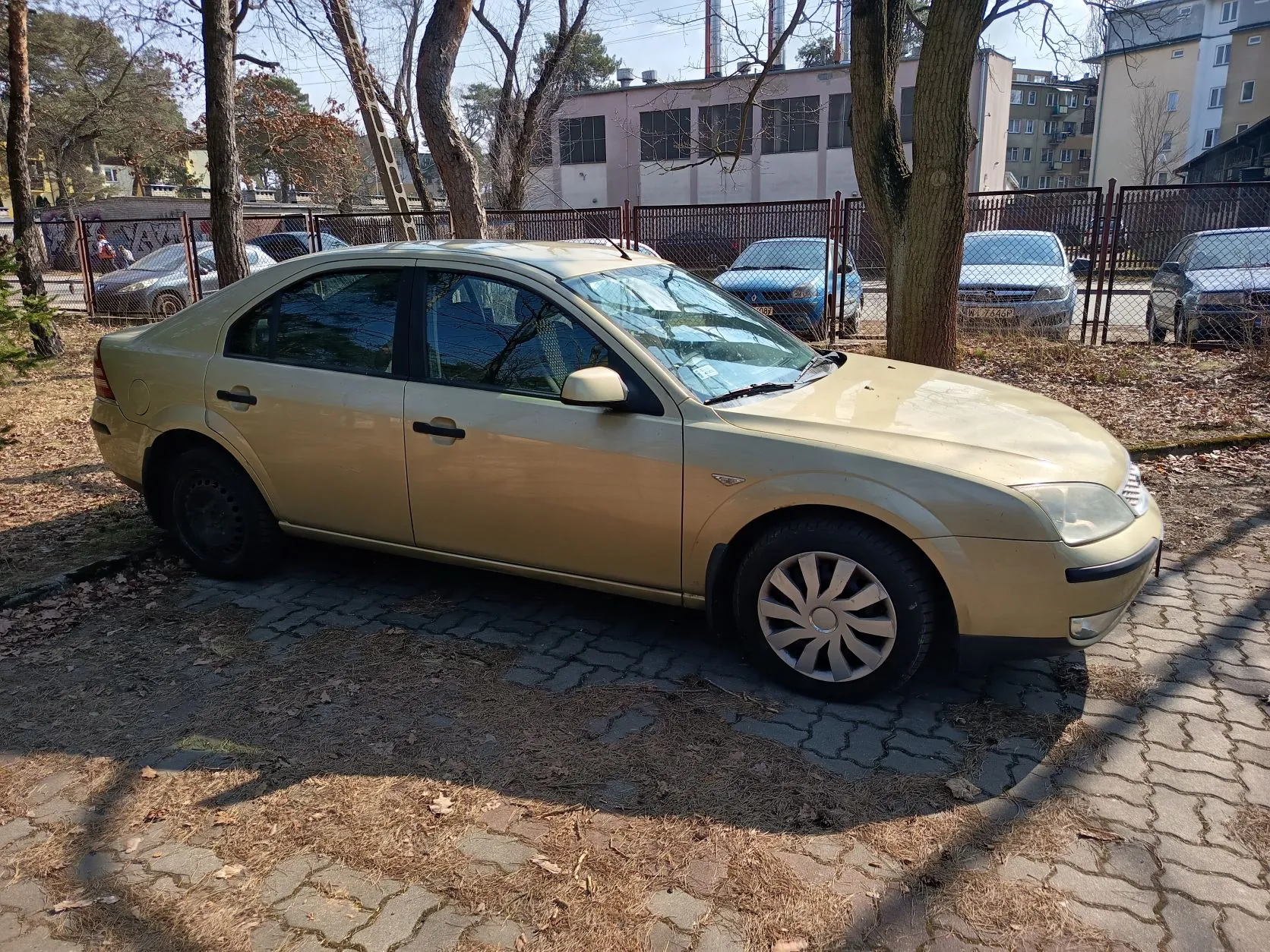 Złoty sedan zaparkowany na brukowanej powierzchni. W tle drzewa bez liści, metalowe ogrodzenie, inne zaparkowane samochody oraz budynki mieszkalne i przemysłowe. Na ziemi sucha trawa i opadłe liście. Słoneczna pogoda, cienie wskazują na jasne światło dzienne.