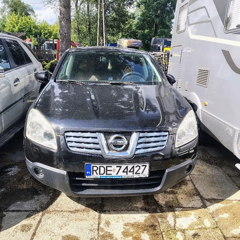 Czarny samochód marki Nissan zaparkowany na kostce brukowej. Widoczny jest przód pojazdu z tablicą rejestracyjną o numerze RDE 74427. Po lewej stronie znajduje się srebrny samochód, a po prawej biały kamper.