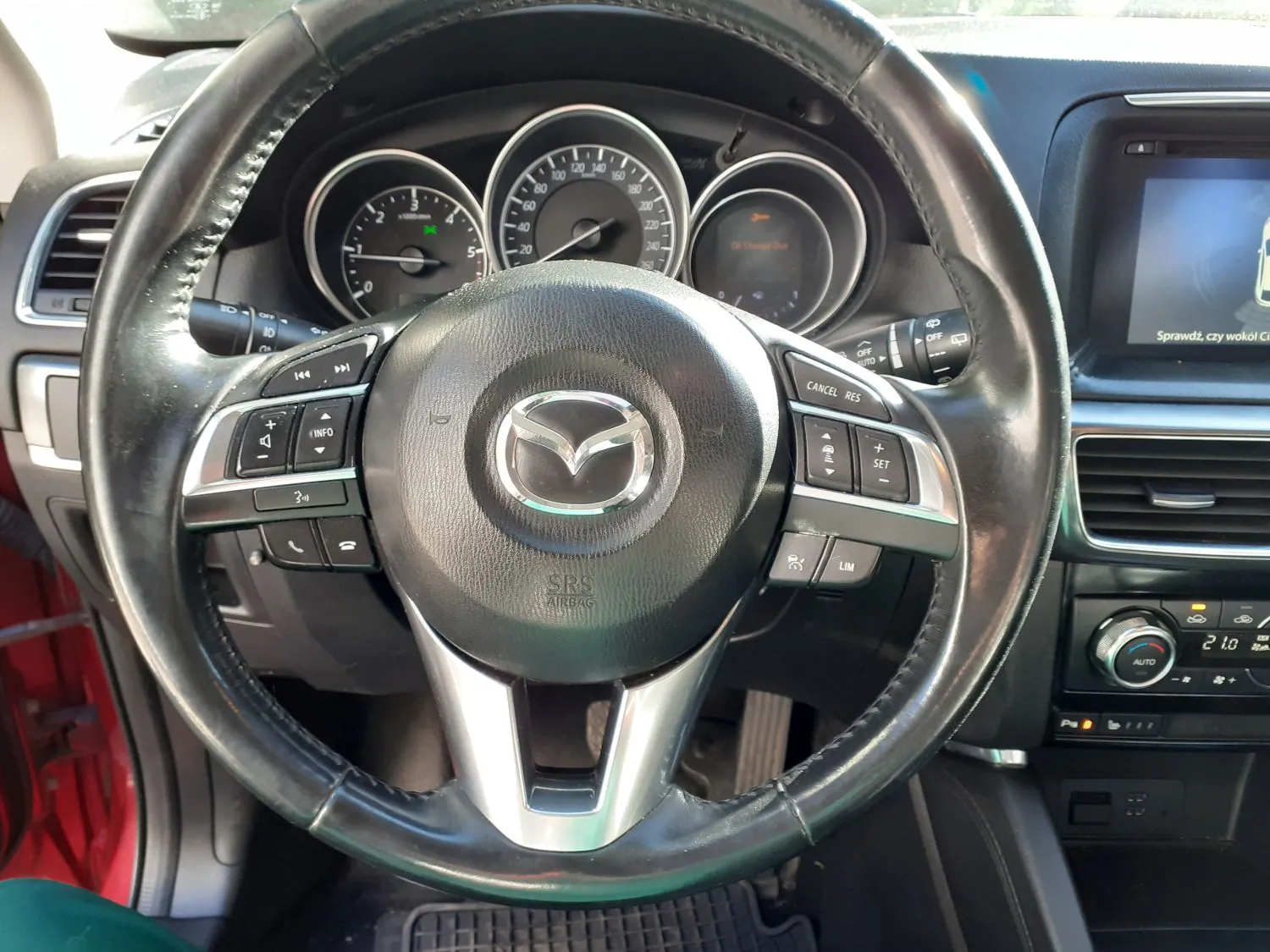 Widok z perspektywy kierowcy na wnętrze samochodu marki Mazda. Na pierwszym planie znajduje się czarna, skórzana kierownica z logo Mazdy w centrum oraz przyciskami sterującymi po obu stronach. W tle widoczna jest deska rozdzielcza z trzema analogowymi wskaźnikami (obrotomierzem, prędkościomierzem i wskaźnikiem paliwa/temperatury) oraz ekranem multimedialnym po prawej stronie, na którym widnieje napis 'Sprawdź, czy wokół c...'. Poniżej ekranu znajduje się panel klimatyzacji z cyfrowym wyświetlaczem wskazującym temperaturę 21.0 stopni.