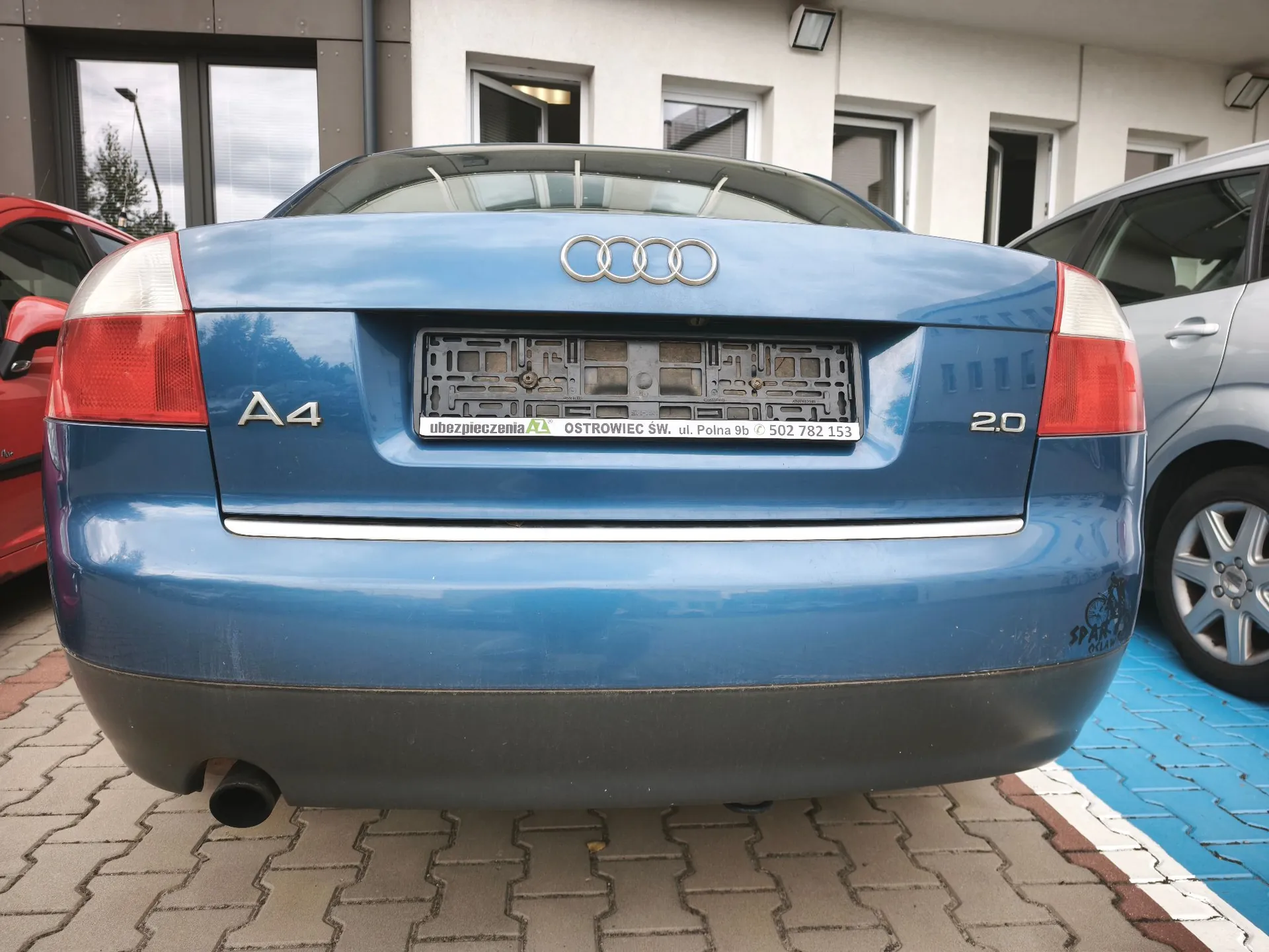 Tylny widok niebieskiego samochodu marki Audi A4 2.0. Pojazd posiada srebrne logo czterech pierścieni na klapie bagażnika oraz oznaczenia modelu 'A4' i silnika '2.0'. W miejscu tablicy rejestracyjnej znajduje się ramka z napisem: 'ubezpieczenia OSTROWIEC SW. ul. Polna 9b 502 782 153'. Samochód zaparkowany jest na kostce brukowej obok czerwonego i srebrnego auta.