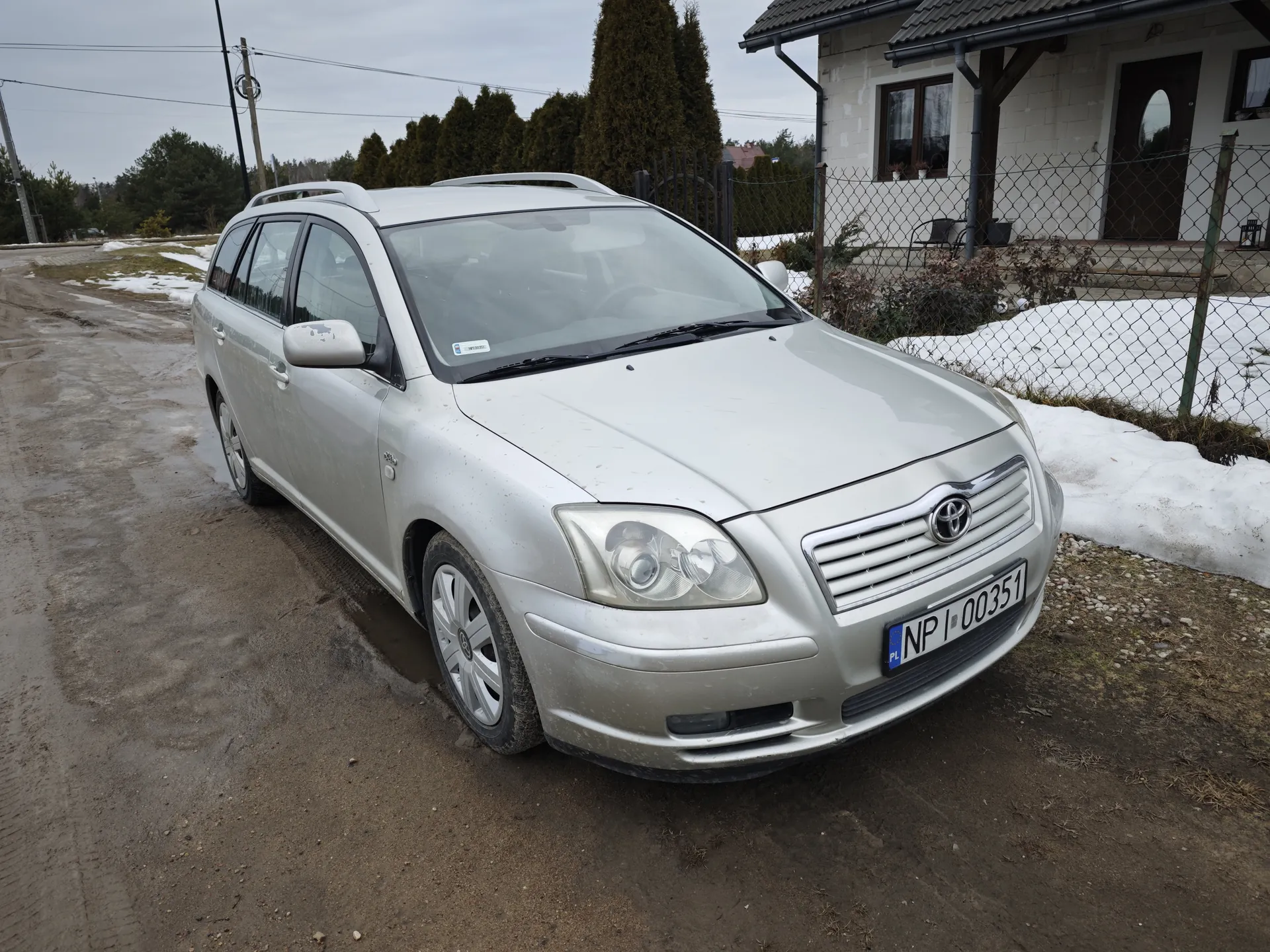 Srebrny samochód osobowy typu kombi marki Toyota zaparkowany na błotnistej drodze. Widoczna tablica rejestracyjna PL NP 00351. W tle budynek mieszkalny z białymi ścianami, ogrodzenie siatkowe, resztki śniegu na ziemi oraz drzewa. Droga pokryta błotem i kałużami, niebo zachmurzone.