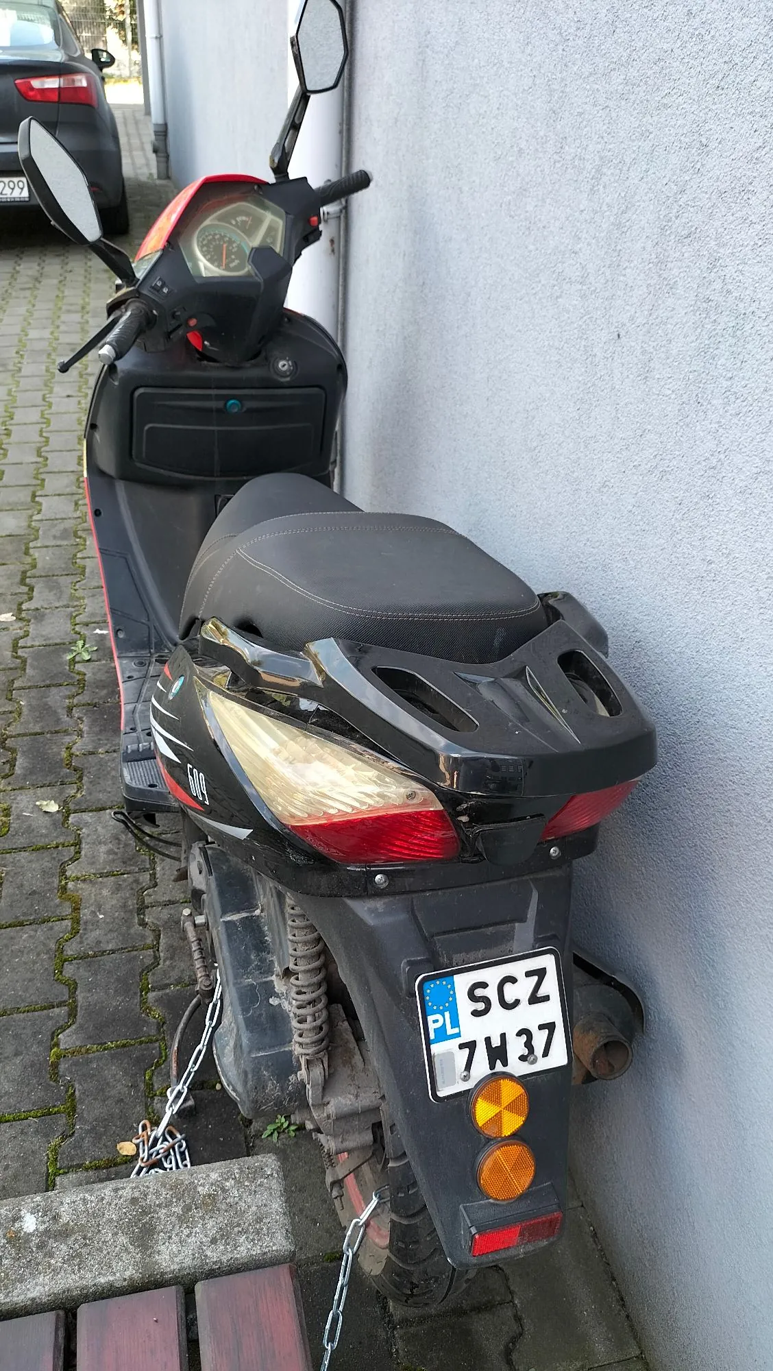 Czarny skuter zaparkowany przy szarej ścianie budynku na kostce brukowej. Widoczny jest tył pojazdu z tablicą rejestracyjną o numerze SCZ 7W37 z oznaczeniem PL. Skuter jest przypięty łańcuchem do elementu konstrukcyjnego. W tle widoczny jest fragment zaparkowanego samochodu.