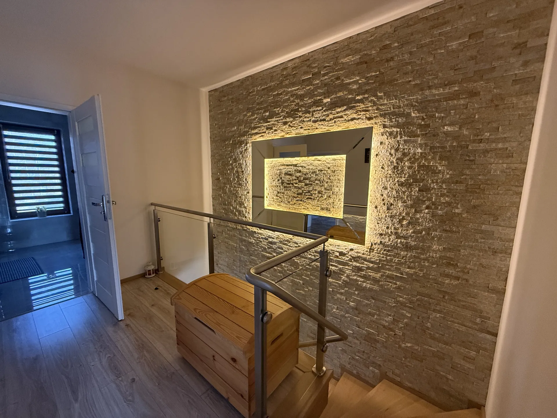 Wnętrze domu z drewnianą podłogą, schodami z metalowym poręczem i szklanymi panelami. Przy schodach znajduje się drewniany kufer. Ściana wykończona kamieniem z dużym lustrzany w otoku oświetlenia LED. Po lewej stronie otwarte drzwi prowadzące do pomieszczenia z oknem z poziomymi żaluzjami. Sufit biały, oświetlenie naturalne i sztuczne.
