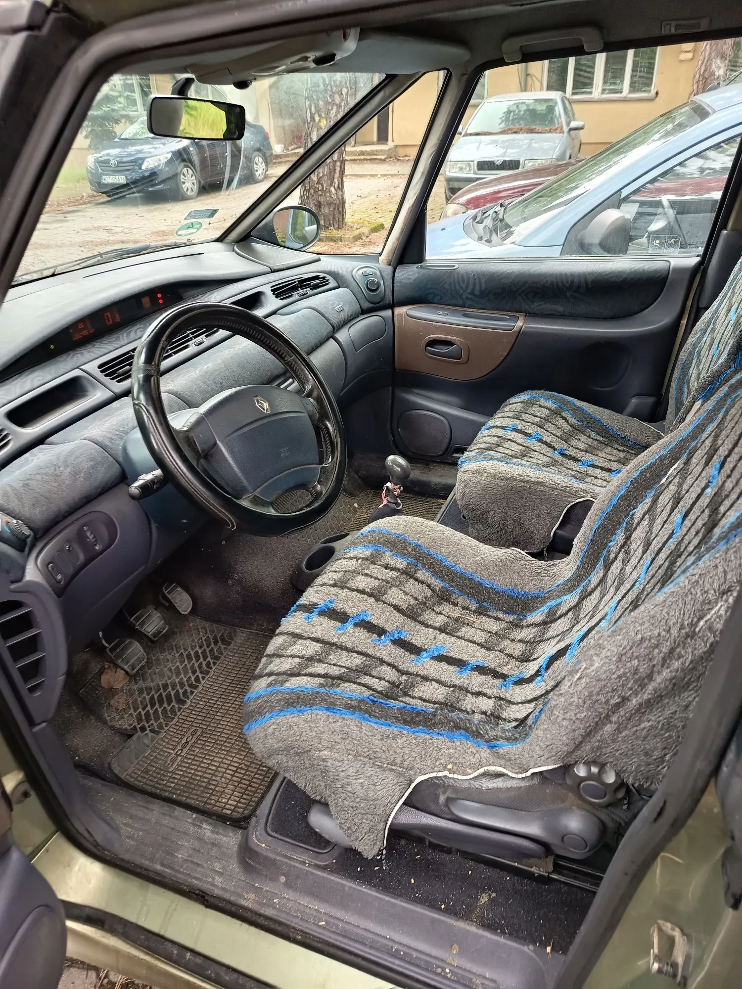 Wnętrze samochodu marki Renault widziane z perspektywy otwartych drzwi pasażera. Widoczna jest kierownica z logo Renault, deska rozdzielcza, pedały oraz fotel kierowcy przykryty szarym kocem w poziome pasy z niebieskimi akcentami. Na podłodze znajduje się gumowa mata. Przez przednią szybę i okna widoczne są inne zaparkowane samochody oraz fragmenty budynków.
