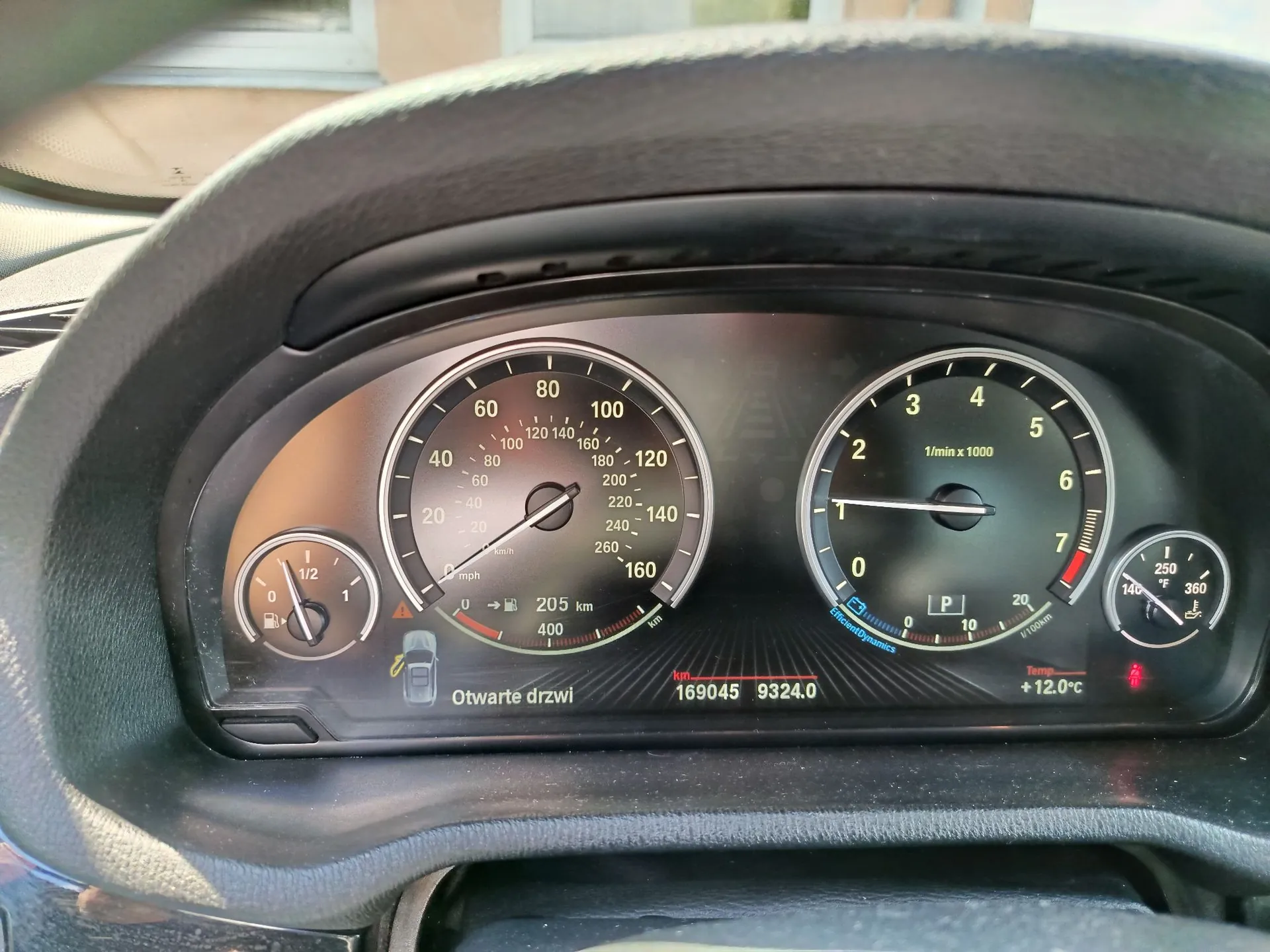Widok z perspektywy kierowcy na deskę rozdzielczą samochodu. W centrum znajduje się prędkościomierz z maksymalną wartością 260 km/h, po prawej obrotomierz z oznaczeniem 1/min x 1000, a po lewej wskaźnik temperatury paliwa. Na wyświetlaczu cyfrowym widnieje napis „Otwarte drzwi”, przebieg 169045 km oraz temperatura zewnętrzna +12.0°C. Widoczny jest również wskaźnik poziomu paliwa oraz ikona otwartych drzwi samochodu.