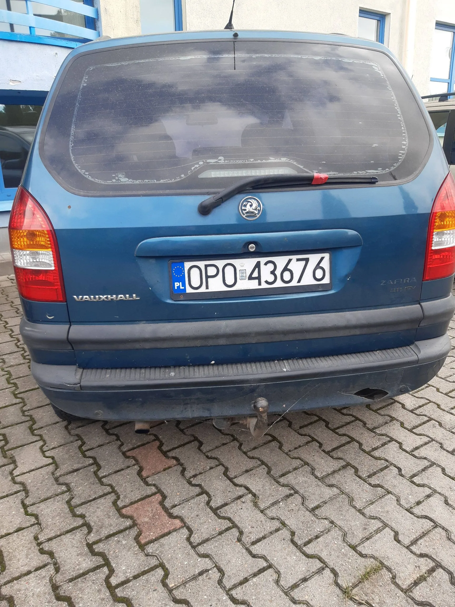 Tylny widok niebieskiego samochodu marki Vauxhall Zafira. Pojazd posiada polską tablicę rejestracyjną o numerze OPO 43676. Na klapie bagażnika widoczne są napisy 'VAUXHALL' po lewej stronie oraz 'ZAFIRA' po prawej. Samochód stoi na kostce brukowej.