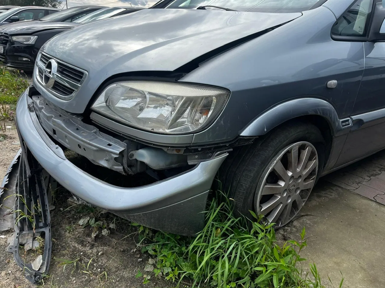 Zbliżenie na przód uszkodzonego, srebrno-niebieskiego samochodu osobowej marki Opel. Przedni zderzak jest częściowo oderwany i zwisa z lewej strony, odsłaniając wewnętrzne elementy konstrukcyjne. Maska samochodu jest lekko podniesiona i odkształcona w centralnej części. Widoczny jest prawy reflektor oraz logo marki na grillu.