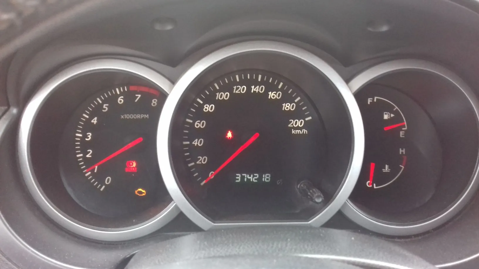 Zdjęcie deski rozdzielczej samochodu z trzema okrągłymi wskaźnikami. Lewy wskaźnik to obrotomierz z oznaczeniem x1000RPM i skalą do 8. Środkowy, największy wskaźnik to prędkościomierz z prędkością w km/h, skalą do 200 oraz cyfrowym licznikiem przebiegu wyświetlającym wartość 374218. Prawy wskaźnik pokazuje poziom paliwa (skala F-E) i temperaturę silnika (skala H-C). Na panelu świecą się czerwone kontrolki ostrzegawcze oraz pomarańczowa kontrolka silnika.