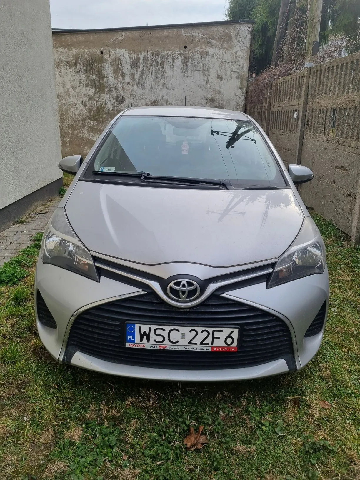 Srebrny samochód marki Toyota z tablicą rejestracyjną WSC 22F6 zaparkowany na trawie obok betonowej ściany i drewnianego ogrodzenia. Na tablicy widoczne są dodatkowe oznaczenia, w tym 'PL' oraz logo marki Toyota na masce. W tle znajdują się drzewa i chmurne niebo.