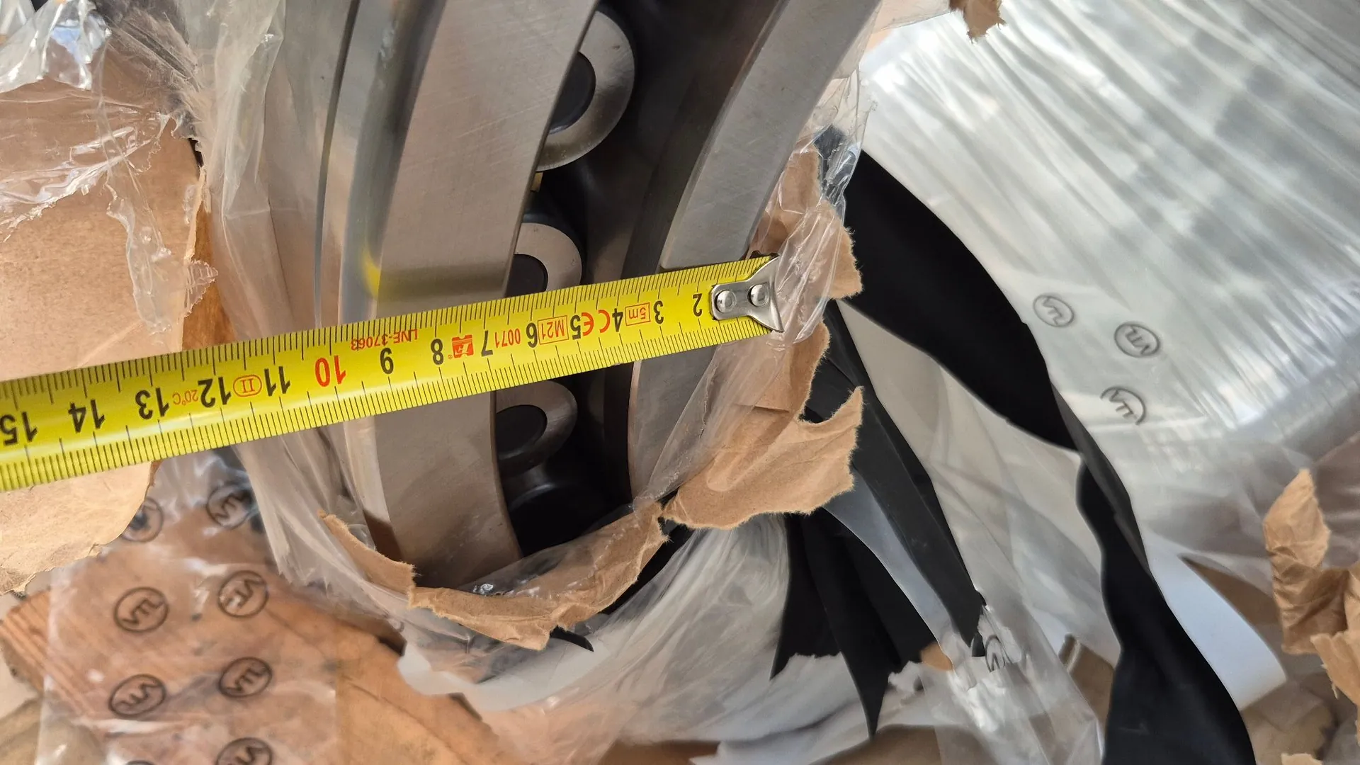 Zbliżenie na metalowy element mechaniczny, prawdopodobnie łożysko toczne, owinięte w folię ochronną i papier pakowy. Do pomiaru średnicy elementu użyta jest żółta miara zwijana, na której widoczne są oznaczenia centymetrów od 2 do 15.