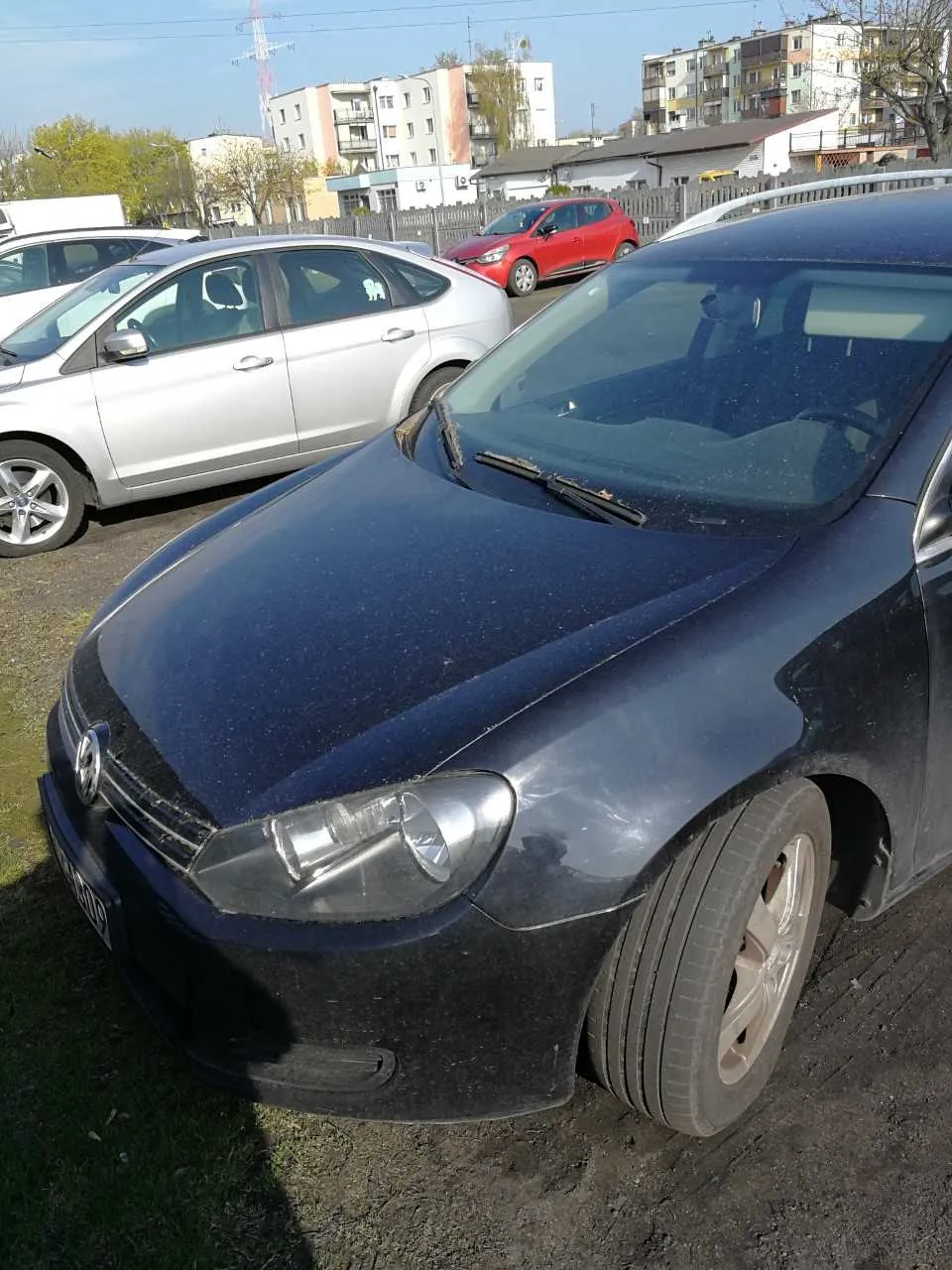 Ciemnoniebieski samochód marki Volkswagen na pierwszym planie, zaparkowany na podłożu ziemnym. W tle srebrny i czerwony samochód, budynki mieszkalne, ogrodzenie oraz drzewa. Niebo jest jasne, pogoda słoneczna. Samochody są ustawione pionowo, horyzont jest poziomy.