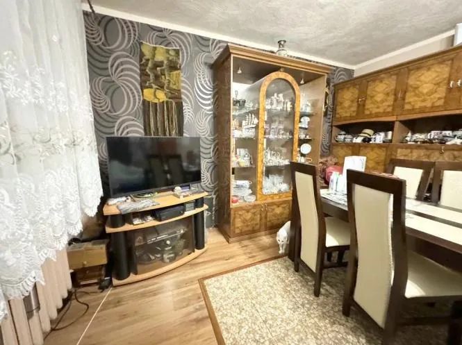 Wnętrze pomieszczenia z drewnianą podłogą, na której znajduje się dywan. Po lewej stronie białe koronkowe zasłony i telewizor na drewnianym stoliku z półkami. Za telewizorem tapeta z geometrycznym wzorem i obrazem. W centrum szafka z szklanymi drzwiami wypełniona porcelaną i szklanymi przedmiotami. Po prawej stronie stół z krzesłami o beżowych oparciach oraz wiszące szafki z dekoracjami. Sufit z matowym wykończeniem.