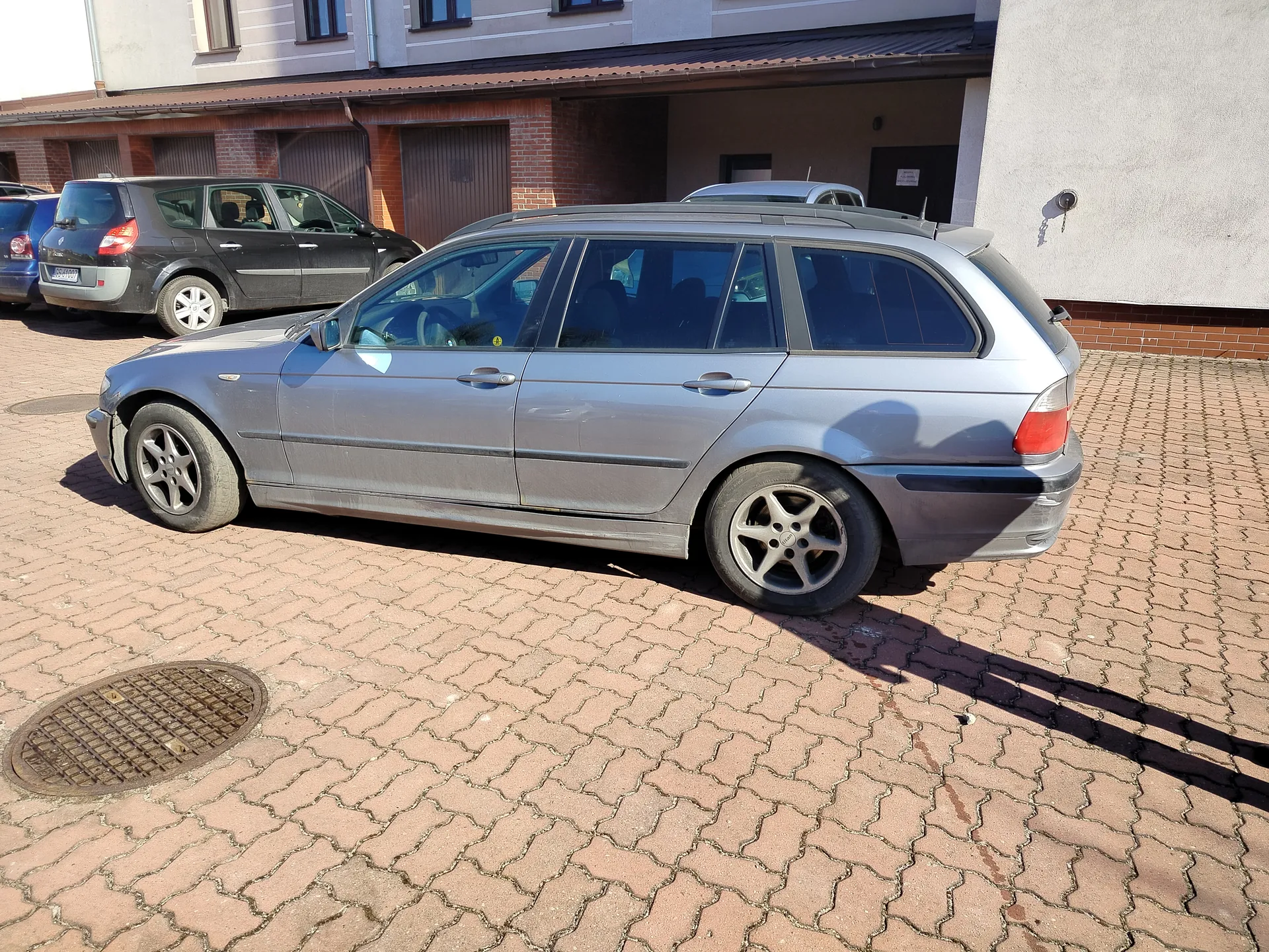 Srebrny samochód typu kombi zaparkowany na brukowanym placu. W tle widoczne są inne pojazdy: ciemnoszary samochód osobowy i niebieski samochód. Za nimi budynek z ceglaną elewacją, bramami garażowymi oraz oknami na piętrze. Na pierwszym planie pokrywa studzienki kanalizacyjnej. Cienie wskazują na słoneczny dzień.