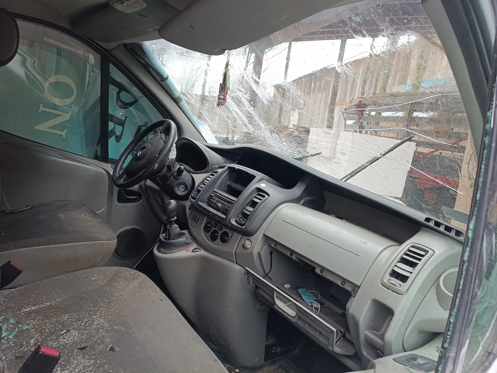 Wnętrze samochodu z widoczną kierownicą po lewej stronie, zniszczonym przednim szybą z pęknięciami, otwartą klapką bocznej konsoli i niebieską maską wewnątrz. Na przedniej szybie widoczny wiszący przedmiot. W tle za szybą drewniana konstrukcja i budynki. Na bocznej szybie widoczny napis NO. Siedzenia są zabrudzone, a deska rozdzielcza w kolorze szarym.