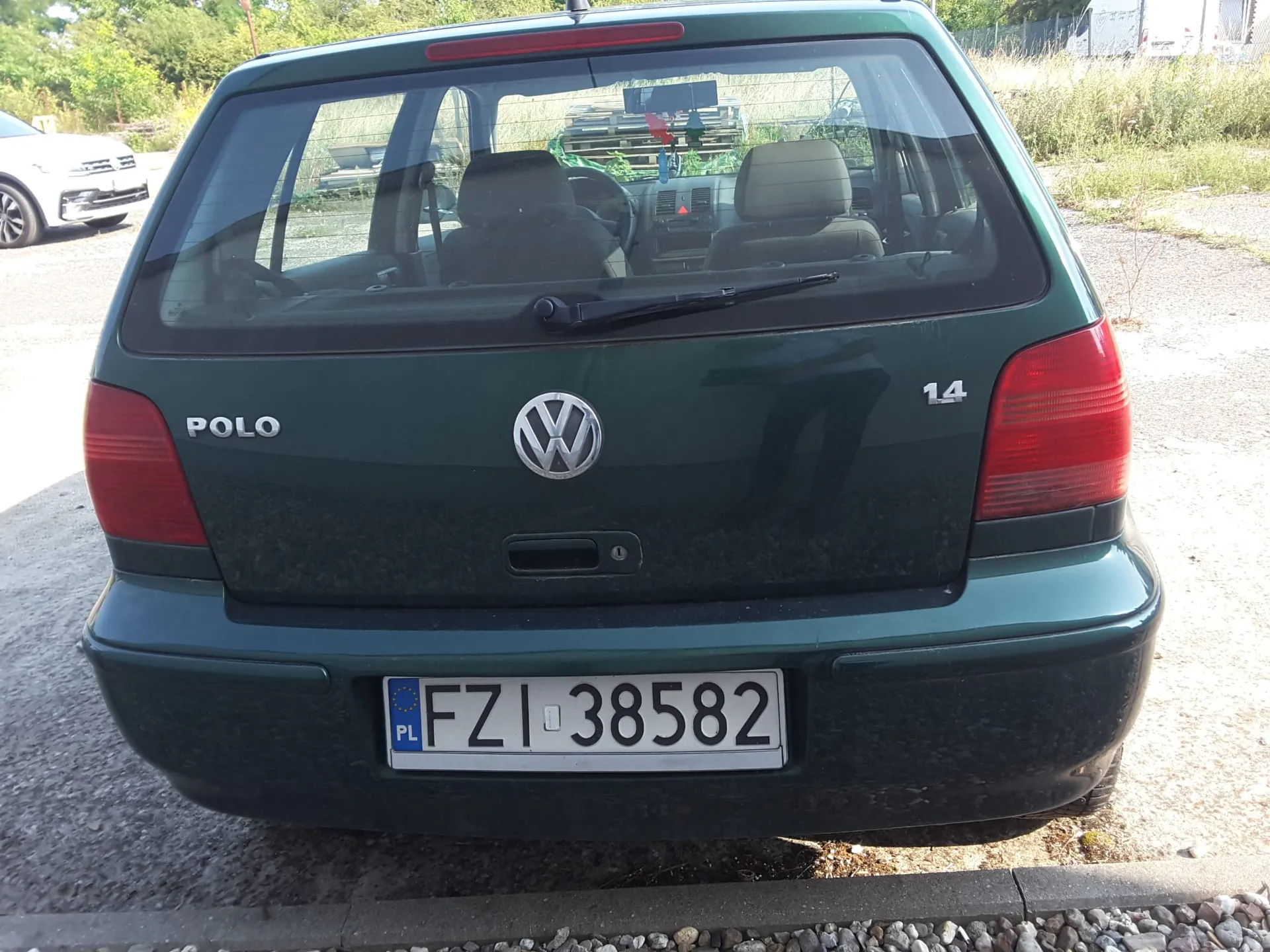 Tylny widok ciemnozielonego samochodu Volkswagen Polo. Na klapie bagażnika widnieje napis 'POLO' po lewej stronie, logo Volkswagena w centrum oraz oznaczenie '14' po prawej stronie. Samochód posiada tablicę rejestracyjną o numerze 'FZI 38582' z oznaczeniem kraju 'PL'. W tle widoczny jest fragment białego samochodu oraz roślinność.