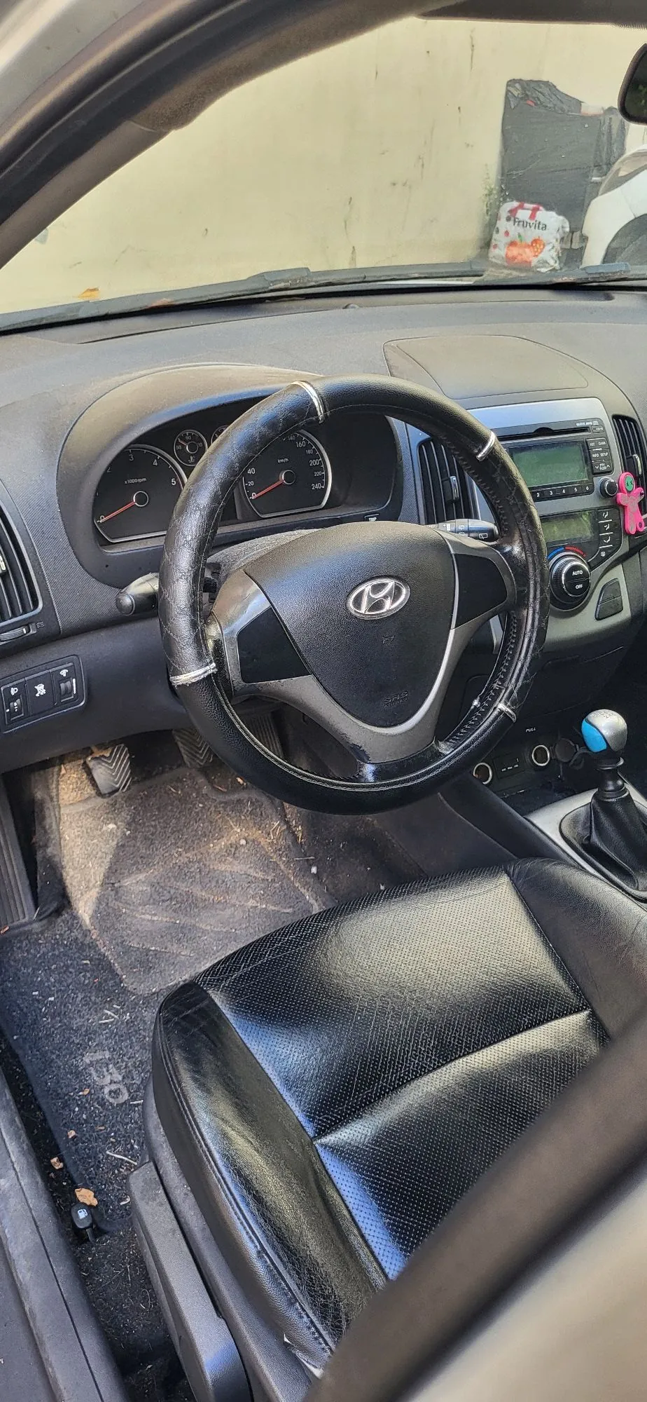 Wnętrze samochodu marki Hyundai z perspektywy kierowcy. Widoczna jest czarna kierownica z okrągłym oplotem, deska rozdzielcza z zegarami, konsola środkowa z radiem i lewarem zmiany biegów z niebieską gałką. W dolnej części zdjęcia znajduje się czarny, skórzany fotel kierowcy oraz dywanik podłogowy.