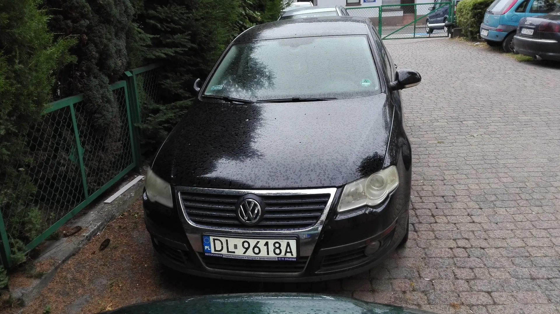 Czarny samochód marki Volkswagen zaparkowany na kostce brukowej. Pojazd jest mokry, prawdopodobnie po deszczu. Widoczna jest tablica rejestracyjna o numerze DL 9618A. Po lewej stronie znajduje się zielony płot i gęste krzewy, a w tle inny niebieski samochód.