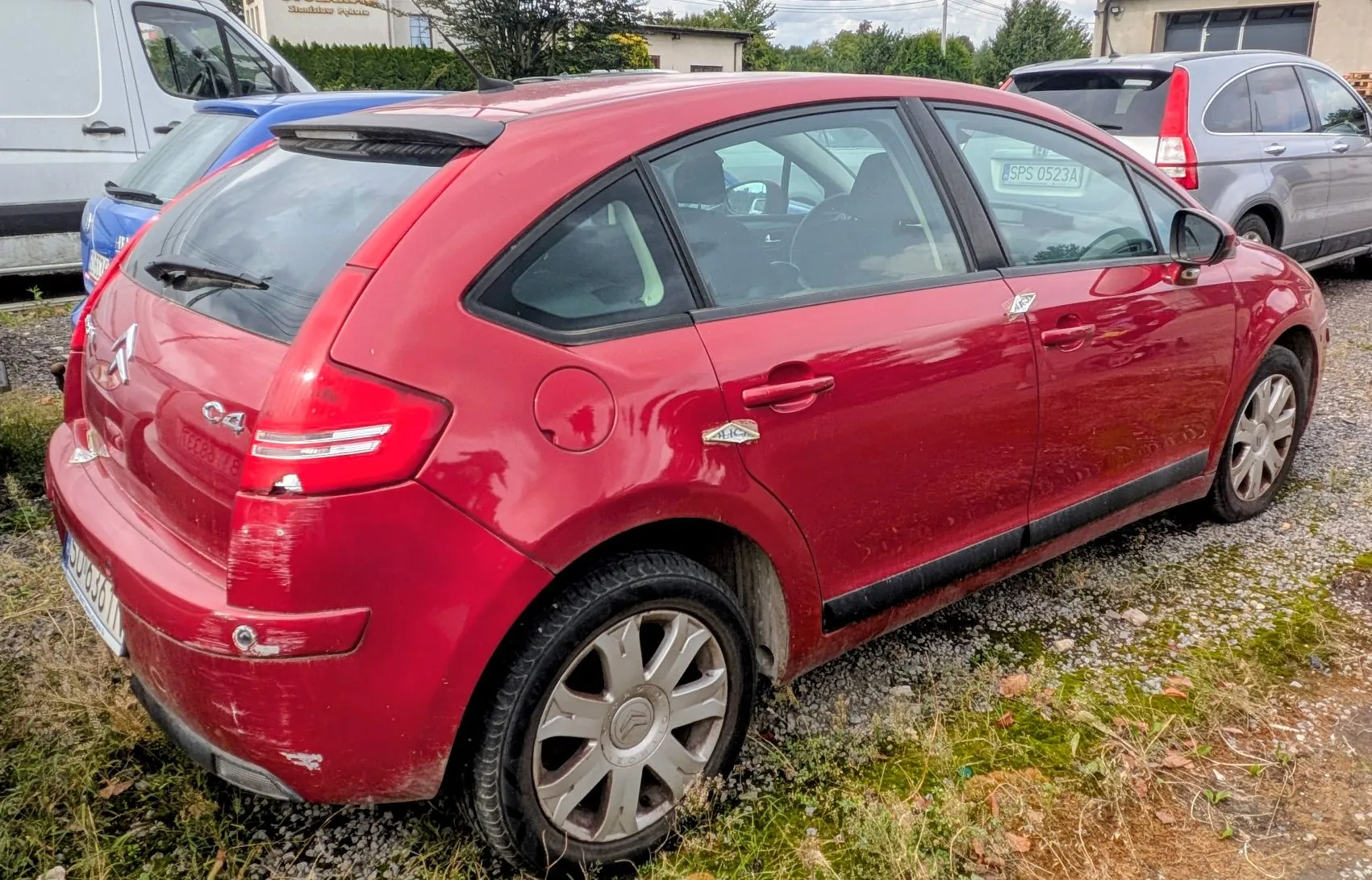 Czerwony samochód marki Citroën C4 w nadwoziu hatchback, zaparkowany na żwirowym podłożu. Pojazd ma widoczne uszkodzenia w postaci zarysowań na tylnym zderzaku i klapie bagażnika. W tle widoczny jest fragment białego vana oraz srebrny samochód z tablicą rejestracyjną SPS 0523A.