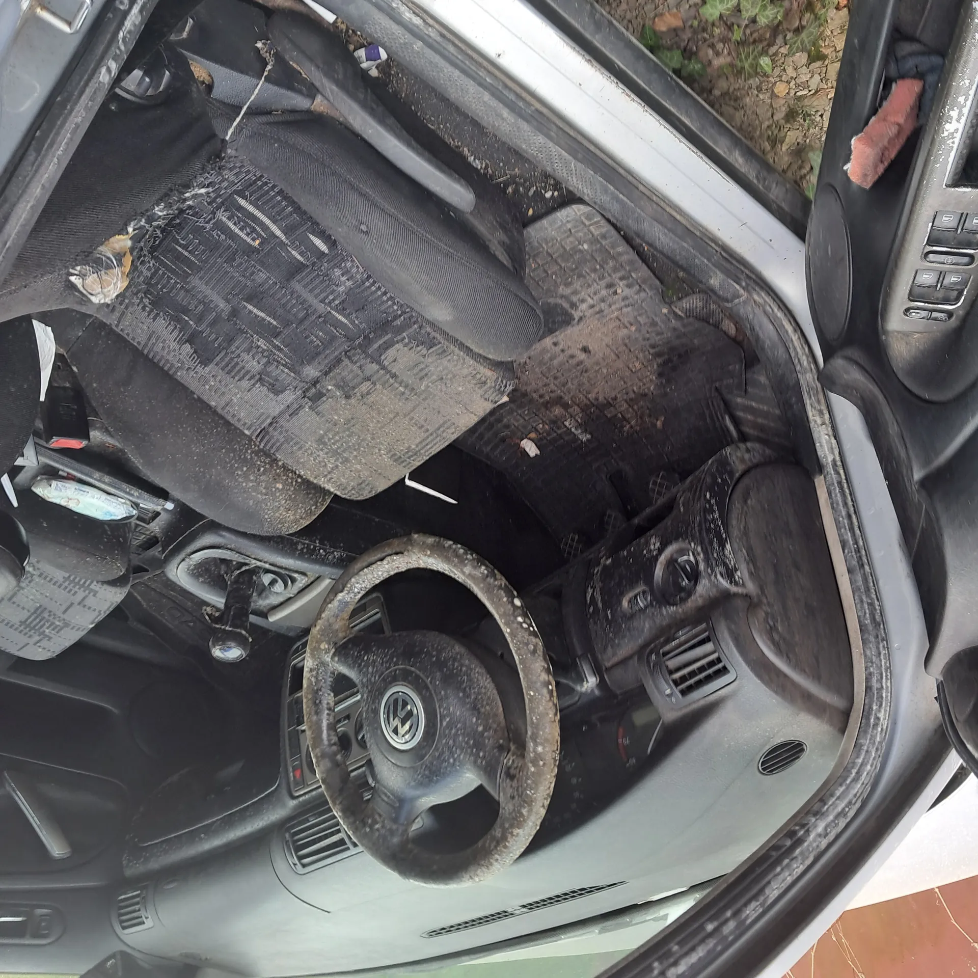 Wnętrze samochodu z widocznym kierownicą marki Volkswagen, zniszczonymi czarnymi siedzeniami, brudnymi dywanikami i deską rozdzielczą. Wnętrze pokryte jest kurzem i śmieciem. Drzwi kierowcy są otwarte, a na zewnątrz widoczny jest fragment ziemi z liśćmi. Na siedzeniu pasażera leżą różne przedmioty, w tym opakowanie.