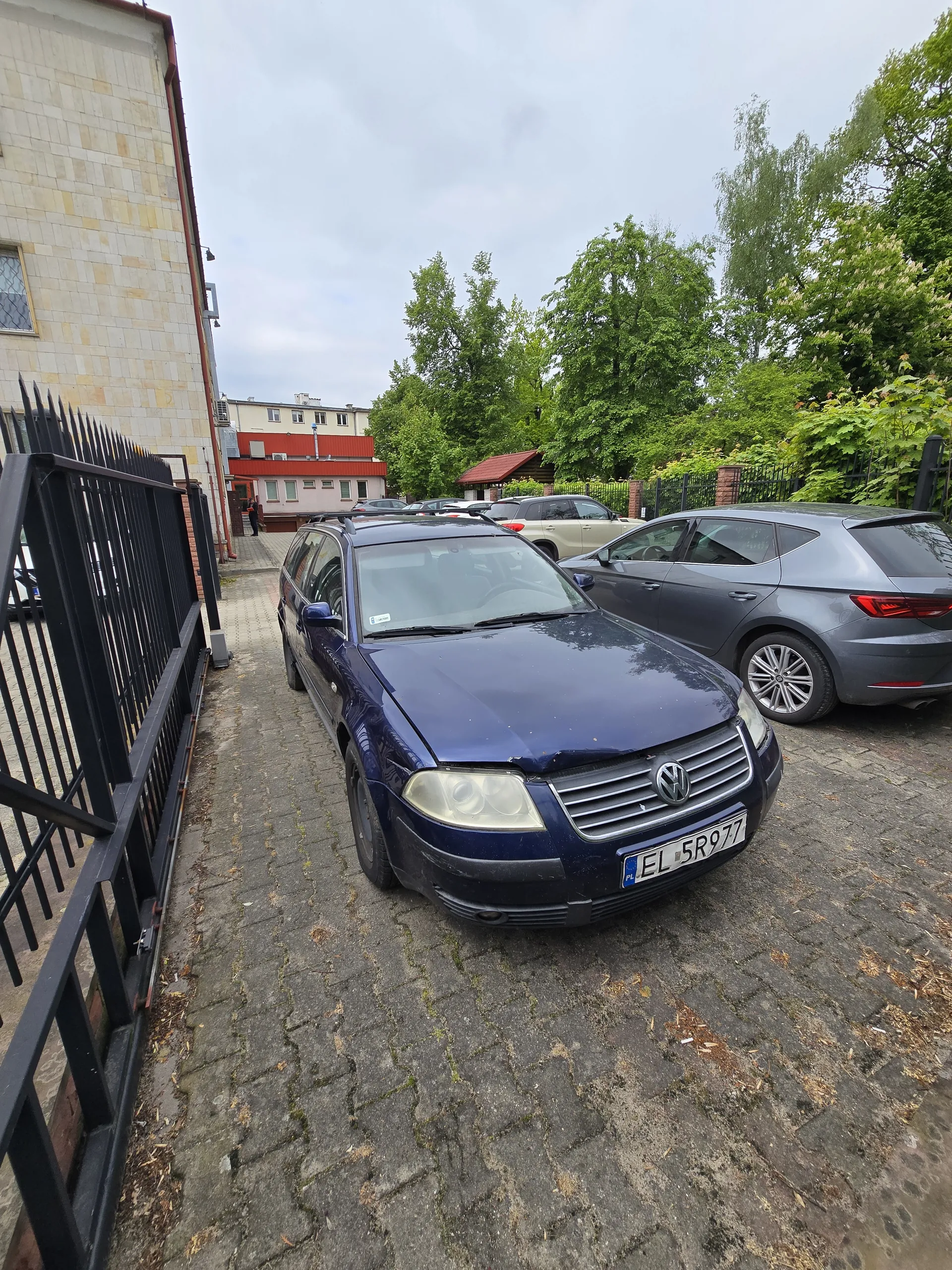 Ciemnoniebieski samochód marki Volkswagen z tablicą rejestracyjną EL 5R97 zaparkowany na brukowanej ulicy. W tle szary samochód, budynki z czerwonymi i jasnymi elewacjami, zielone drzewa oraz chmurne niebo. Widoczna czarna metalowa brama.