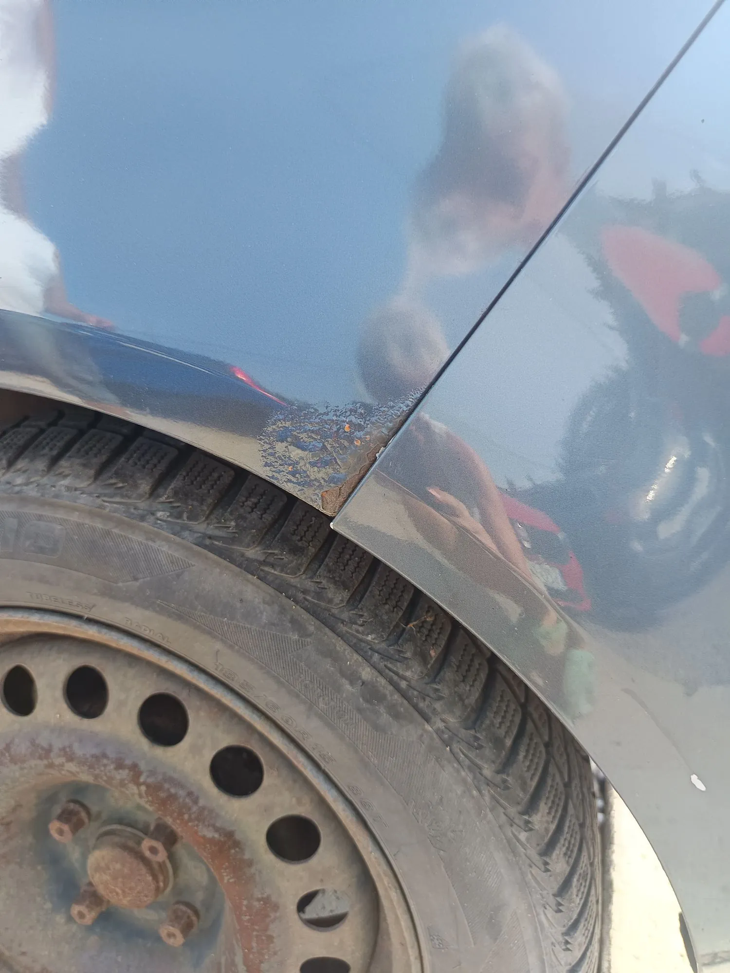 Zbliżenie na fragment nadkola i opony w samochodzie. Lakier samochodu jest w kolorze ciemnoniebieskim i posiada widoczne uszkodzenia, w tym odpryski oraz rdzę w miejscu łączenia elementów karoserii. Poniżej znajduje się czarna opona oraz metalna felga z widocznymi śladami korozji.