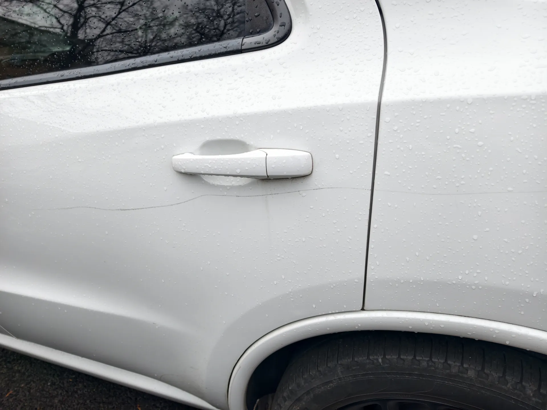 Biały samochód z widocznym fragmentem drzwi, uchwytem drzwiowym i kołem. Na karoserii znajdują się krople deszczu. W tle przez okno widoczne są nagie gałęzie drzew. Samochód stoi na asfaltowej powierzchni.