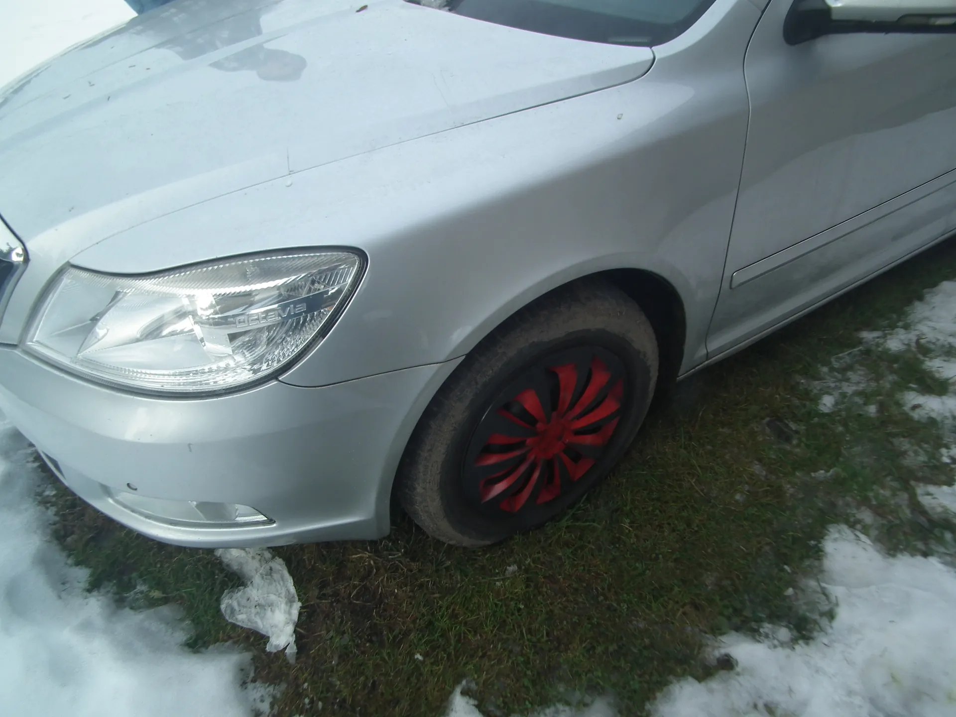 Srebrny samochód marki Octavia, widok przedniej lewej części. Widoczny reflektor z napisem "OCTAVIA", koło z czerwono-czarną felgą. Podwozie na trawie z resztkami śniegu.