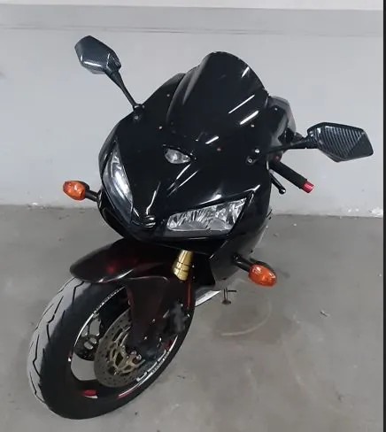 Czarny motocykl sportowy widziany z przodu. Pojazd posiada dwie lusterka w karbonowym wykończeniu, dwa reflektory oraz pomarańczowe kierunkowskazy. Widoczne jest przednie koło z tarczą hamulcową i złotymi amortyzatorami. Motocykl stoi na szarym, betonowym podłożu na tle białej ściany.