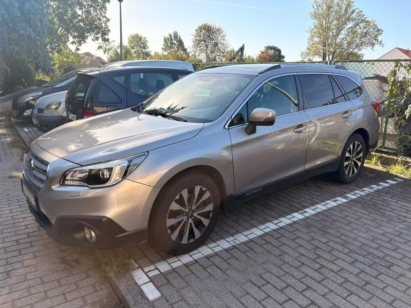 Srebrny samochód marki Subaru Outback zaparkowany na kostce brukowej. Pojazd jest pokazany z przodu i z boku, z widocznym napisem 'OUTBACK' na dolnej listwie bocznej. W tle widoczne są inne zaparkowane samochody, zielone drzewa oraz jasne, bezchmurne niebo.