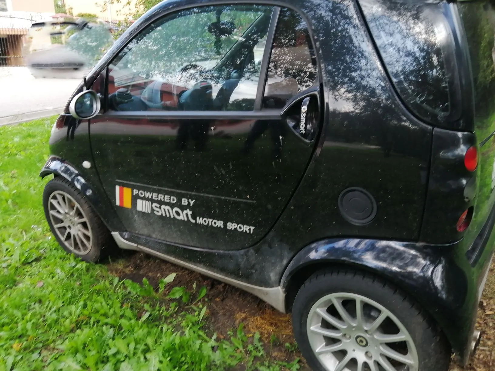 Czarny samochód marki Smart zaparkowany na trawie. Na boku pojazdu znajduje się biały napis: 'POWERED BY Smart MOTOR SPORT' wraz z małą flagą w kolorach żółtym i czerwonym.