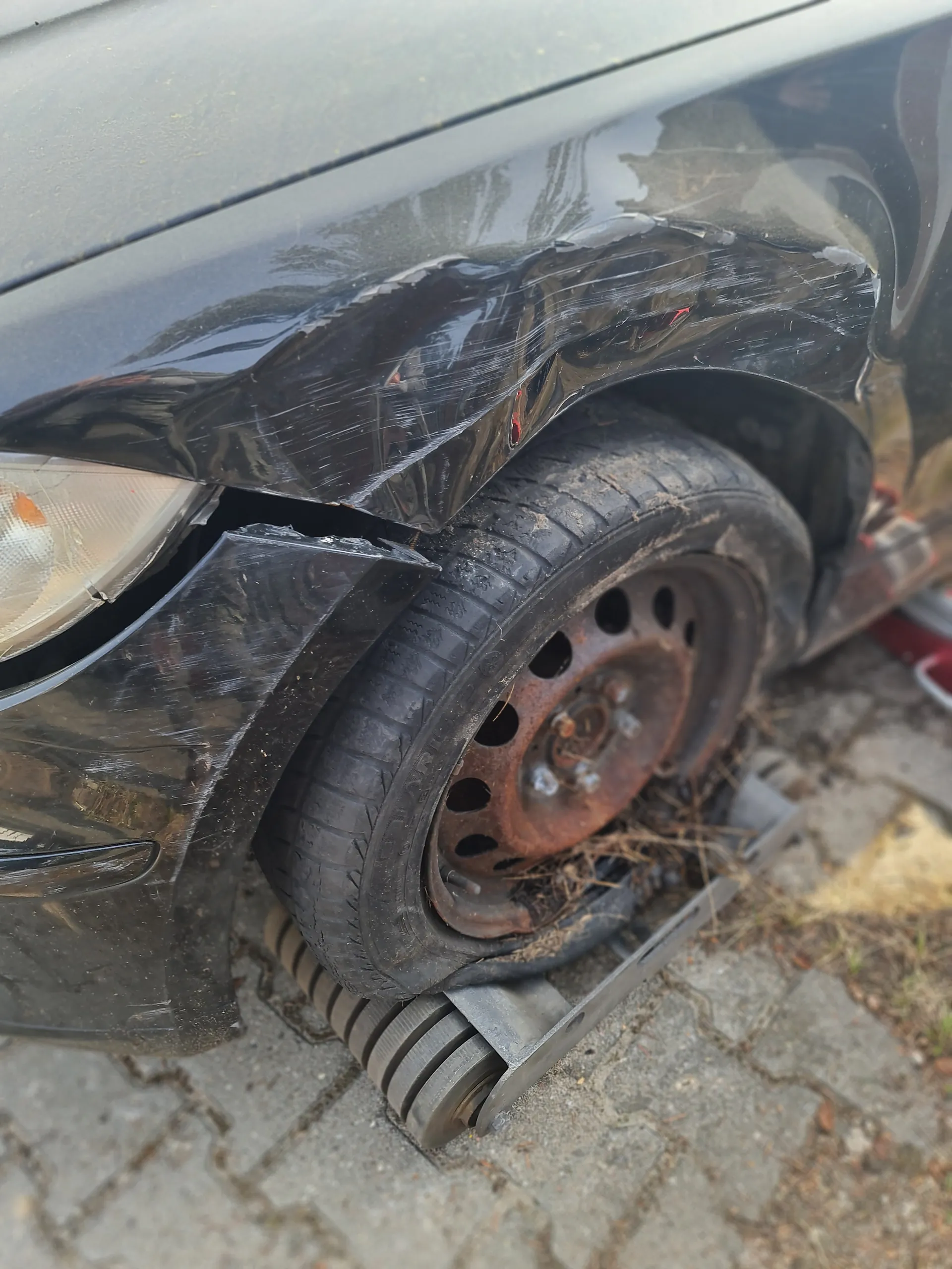 Zbliżenie na uszkodzony przód czarnego samochodu. Widoczna zgnieciona karoseria, pęknięty zderzak, uszkodzony reflektor oraz opona z rdzewiałym felgiem. Samochód znajduje się na brukowanej powierzchni z widocznymi szczelinami i suchą trawą.
