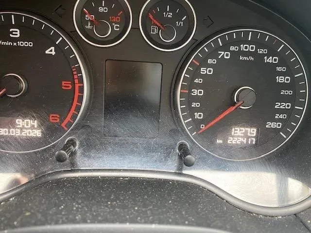 Zbliżenie na deski rozdzielcze samochodu. Po lewej stronie tachometr z zakresem do 6 (1/min x1000), wskazówka blisko 0, cyfrowy wyświetlacz z godziną 9:04 i datą 30.03.2026. W centrum czarny prostokątny ekran. Po prawej prędkościomierz w km/h (zakres do 260), wskazówka na 0, poniżej cyfrowe wartości 13219 km i 2224.17. W górnej części dwa mniejsze wskaźniki: termometr (50-130°C) i wskaźnik paliwa (1/2). Całość na czarnym tle z widocznymi śladami kurzu.