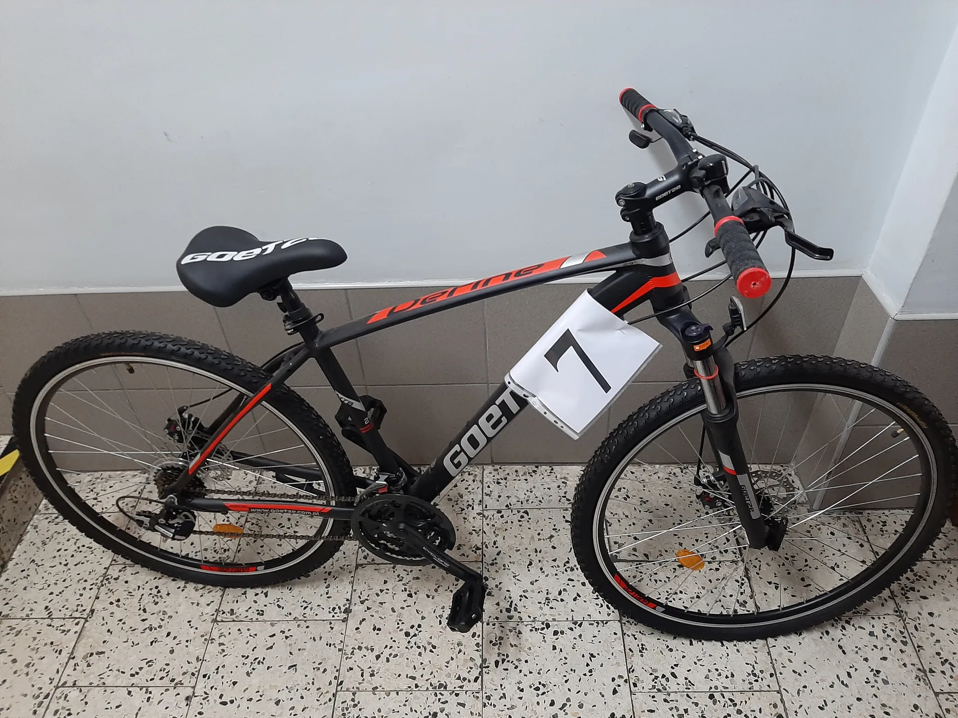 Czarny rower górski z pomarańczowymi akcentami marki GOET, stojący na podłodze z białymi płytkami w kropki. Na ramie roweru widoczny biały znaczek z czarną cyfrą 7. Siedzisko z napisem GOET, kierownica z czerwonymi uchwytami. Tło: szara ściana z brązowymi płytkami na dole i białą częścią na górze.