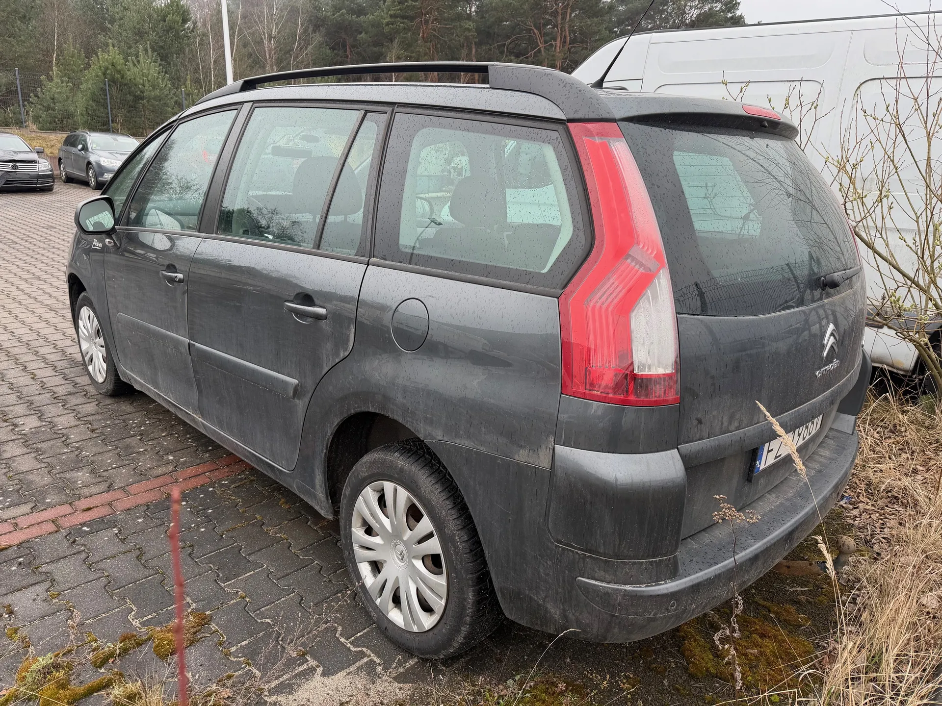 Szary samochód Citroën z brudną karoserią zaparkowany na brukowanym placu. Widoczne są czerwone światła tylnego, srebrne felgi, dachowy bagażnik i tablica rejestracyjna z częściowo widocznym numerem FZ. W tle biały furgon, inne pojazdy, drzewa oraz sucha trawa i mchy na ziemi. Samochód ma cztery drzwi i ciemne szyby.