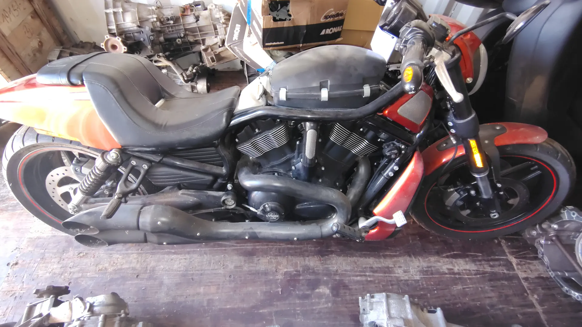 Czerwono-czarny motocykl w warsztacie z czarnym siedzeniem, podwójnymi wydechami i widocznymi elementami silnika. W tle znajdują się mechaniczne części oraz pudełka z napisami 'speedline' i 'ARONAL'. Podłoga warsztatu jest drewniana.