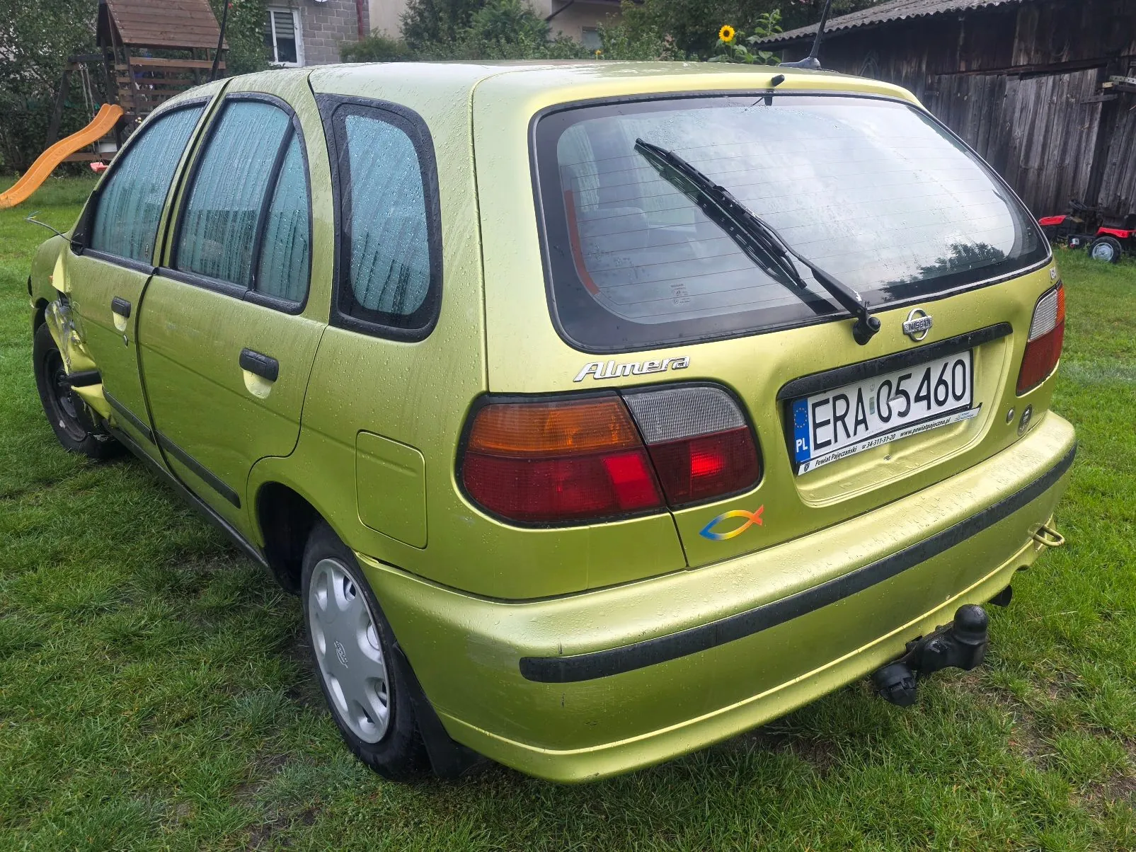 Jasnozielony samochód marki Nissan Almera zaparkowany na trawie. Widoczny jest tył i lewy bok pojazdu. Samochód ma polską tablicę rejestracyjną o numerze ERA 05460. Na klapie bagażnika znajduje się napis 'Almera' oraz logo Nissana. Na tylnym zderzaku zamontowany jest hak holowniczy.