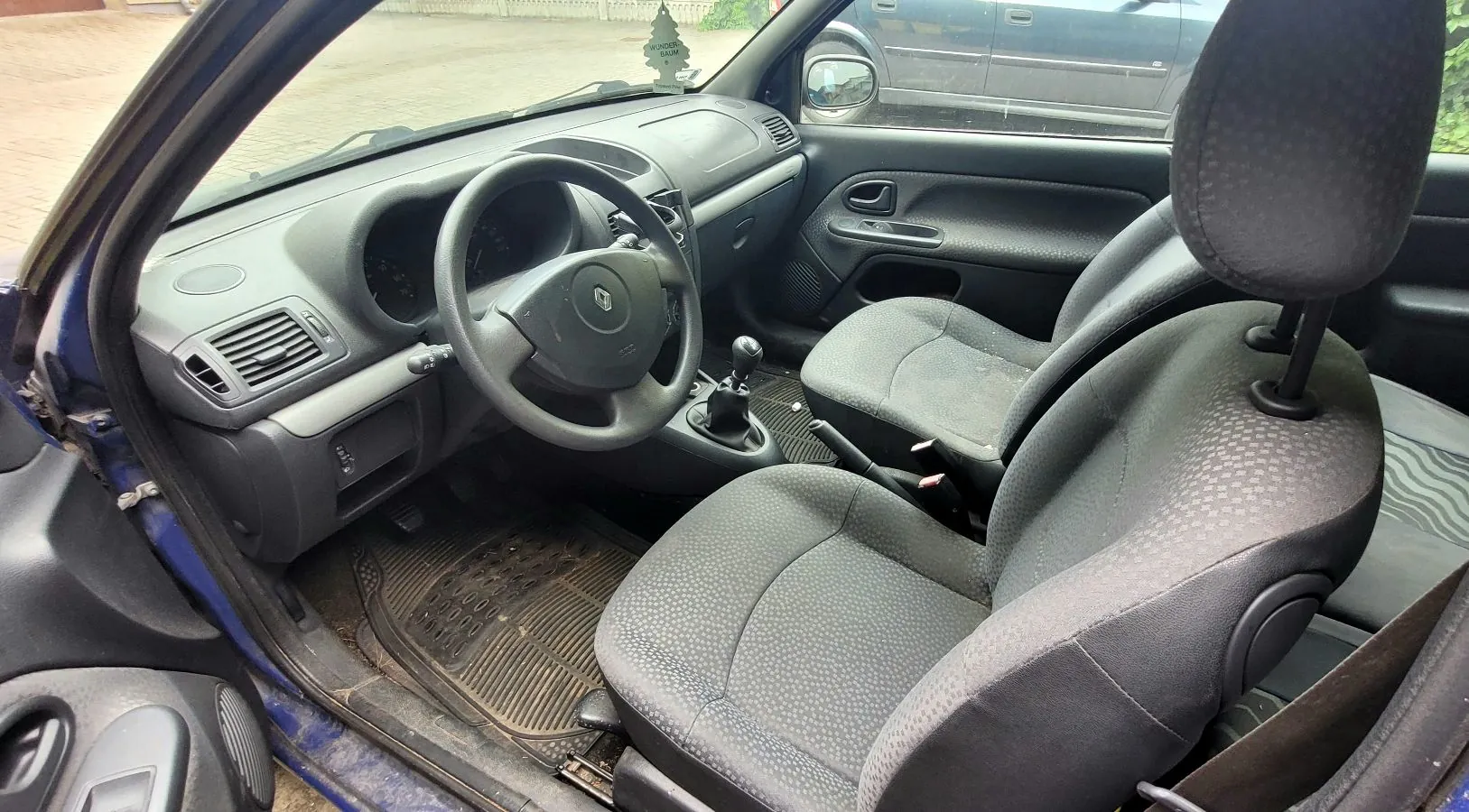 Wnętrze samochodu widziane z perspektywy otwartych przednich drzwi po stronie kierowcy. Widoczna jest czarna deska rozdzielcza, kierownica z logo Renault, dwa przednie fotele z ciemnoszarej tkaniny o drobnej strukturze oraz gumowe dywaniki na podłodze. Przez przednią szybę widoczny jest fragment otoczenia i lusterko boczne.