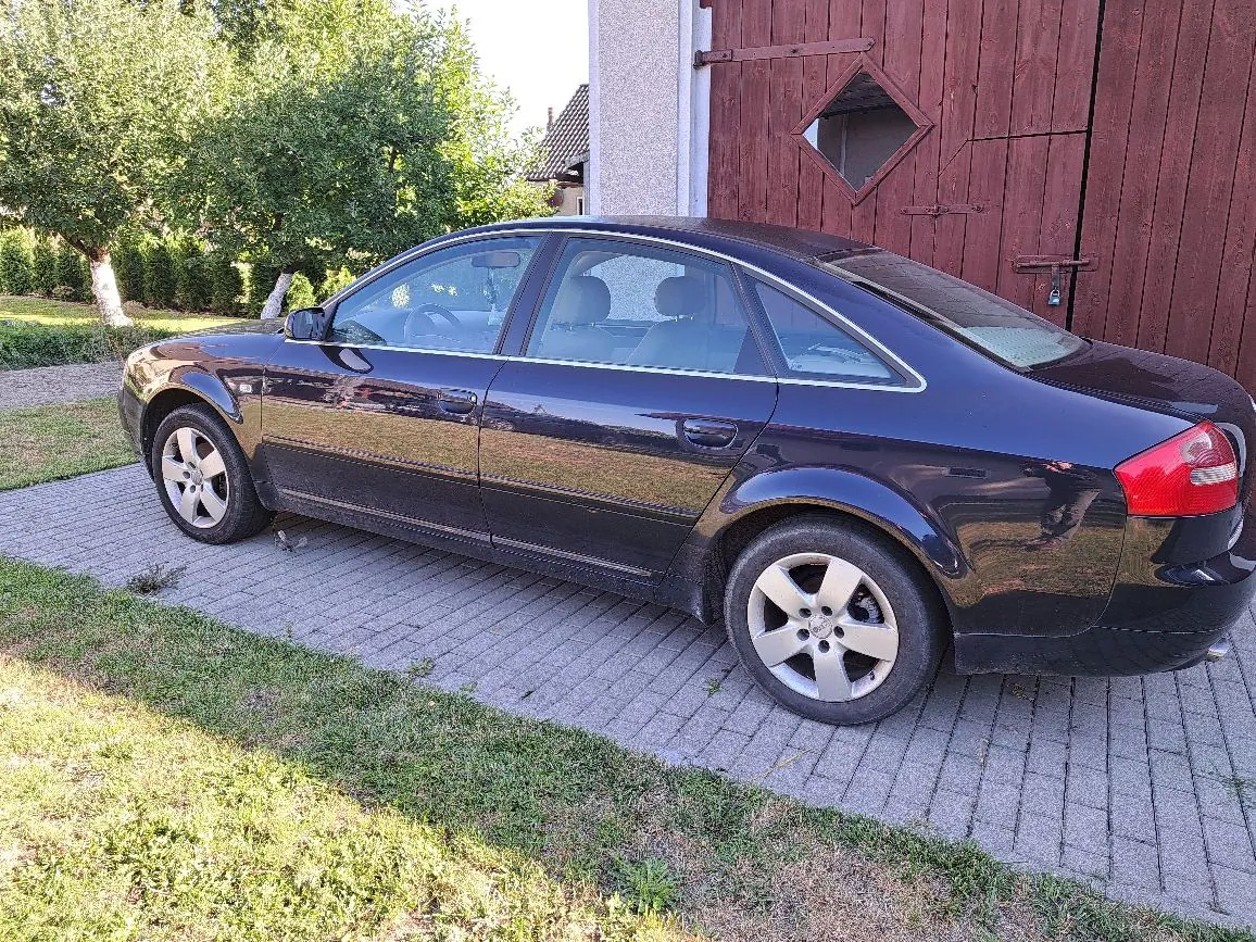 Ciemnofioletowy samochód osobowy typu sedan zaparkowany bokiem na kostce brukowej przed drewnianymi drzwiami garażowymi. W tle widoczne są zielone drzewa i trawnik.