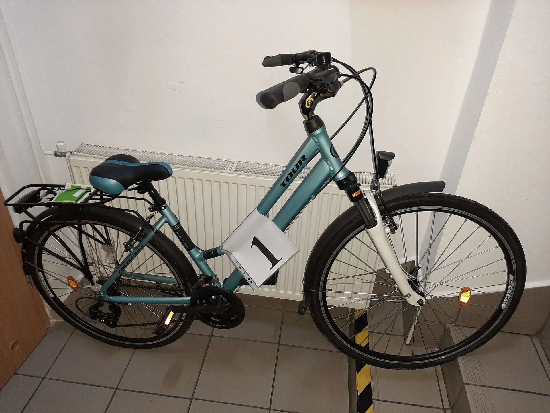 Niebiesko-zielony rower marki TOUR z białą tabliczką z numerem 1 na ramie. Rower ma czarno-niebieskie siedzenie, bagażnik tylny z zielonym przedmiotem, czarne kierownice i przedni błotnik. Znajduje się na tle białego grzejnika, białej ściany i podłogi z szarymi płytkami. Widoczne są również pomarańczowe odblaski na kołach oraz czarno-żółta taśma na podłodze po prawej stronie.