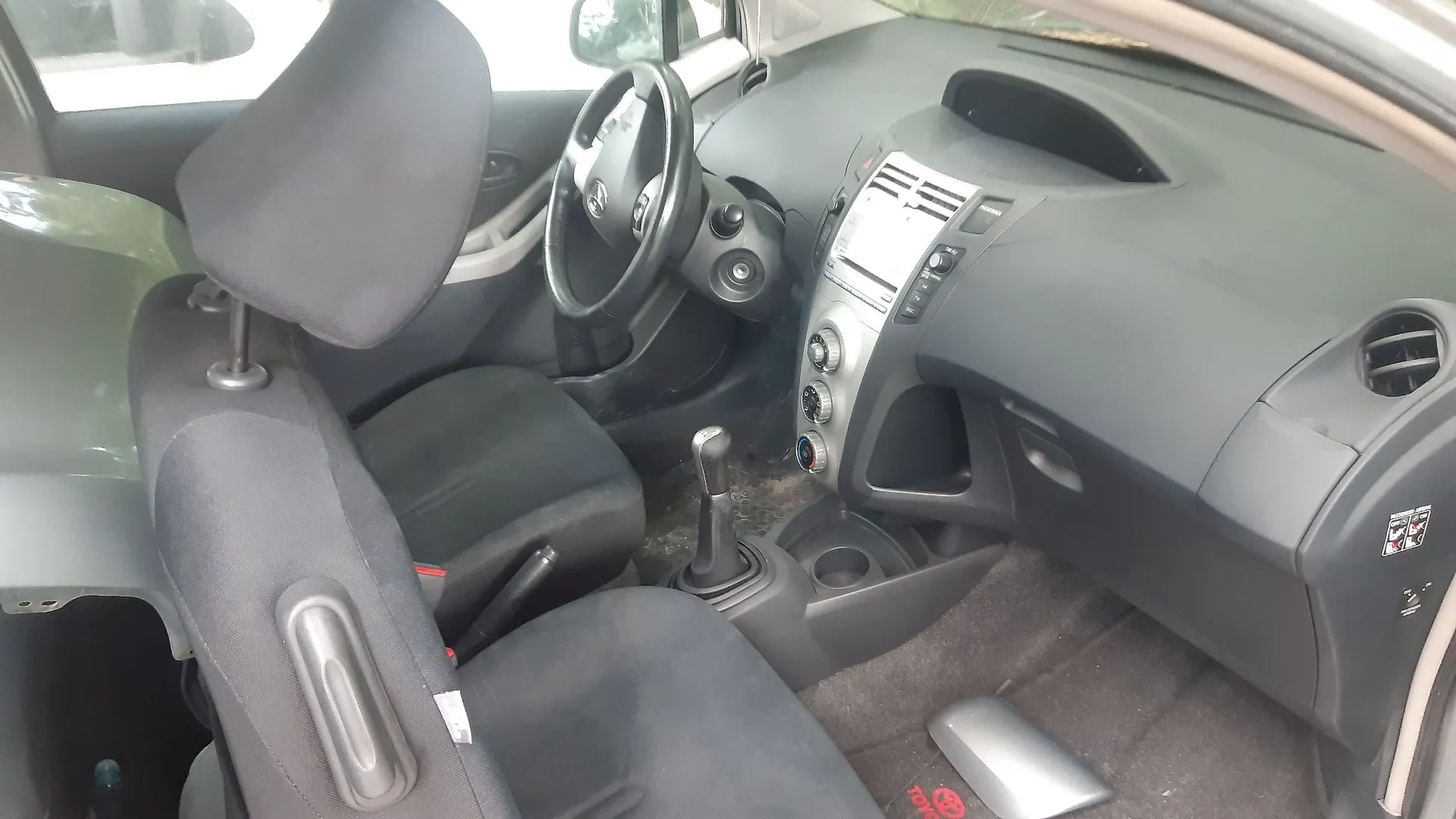 Wnętrze samochodu z perspektywy otwartych drzwi kierowcy. Widoczny jest czarny fotel z zagłówkiem, kierownica z logo Toyoty, deska rozdzielcza z centralnie umieszczonym radiem i pokrętłami klimatyzacji oraz dźwignia zmiany biegów. Na podłodze widoczna jest srebrna stopka z czerwonym napisem Toyota.