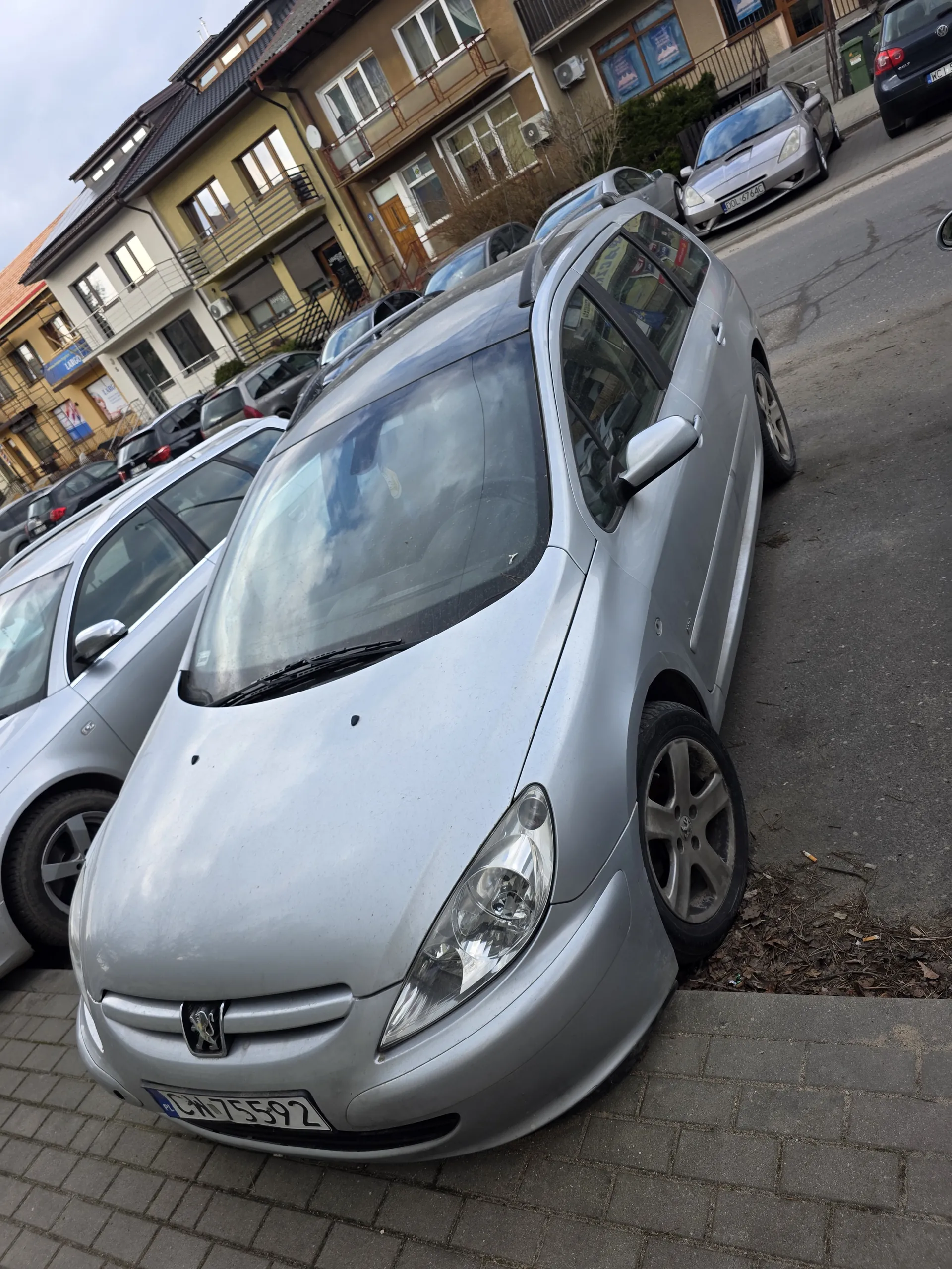 Zdjęcie przedstawia srebrny samochód zaparkowany przy chodniku z ceglanym wykończeniem. Widoczna tablica rejestracyjna CJ 7559L. W tle inne zaparkowane pojazdy oraz wielopiętrowe budynki mieszkalne z oknami i balkonami. Na chodniku znajdują się suche liście i śmieci.