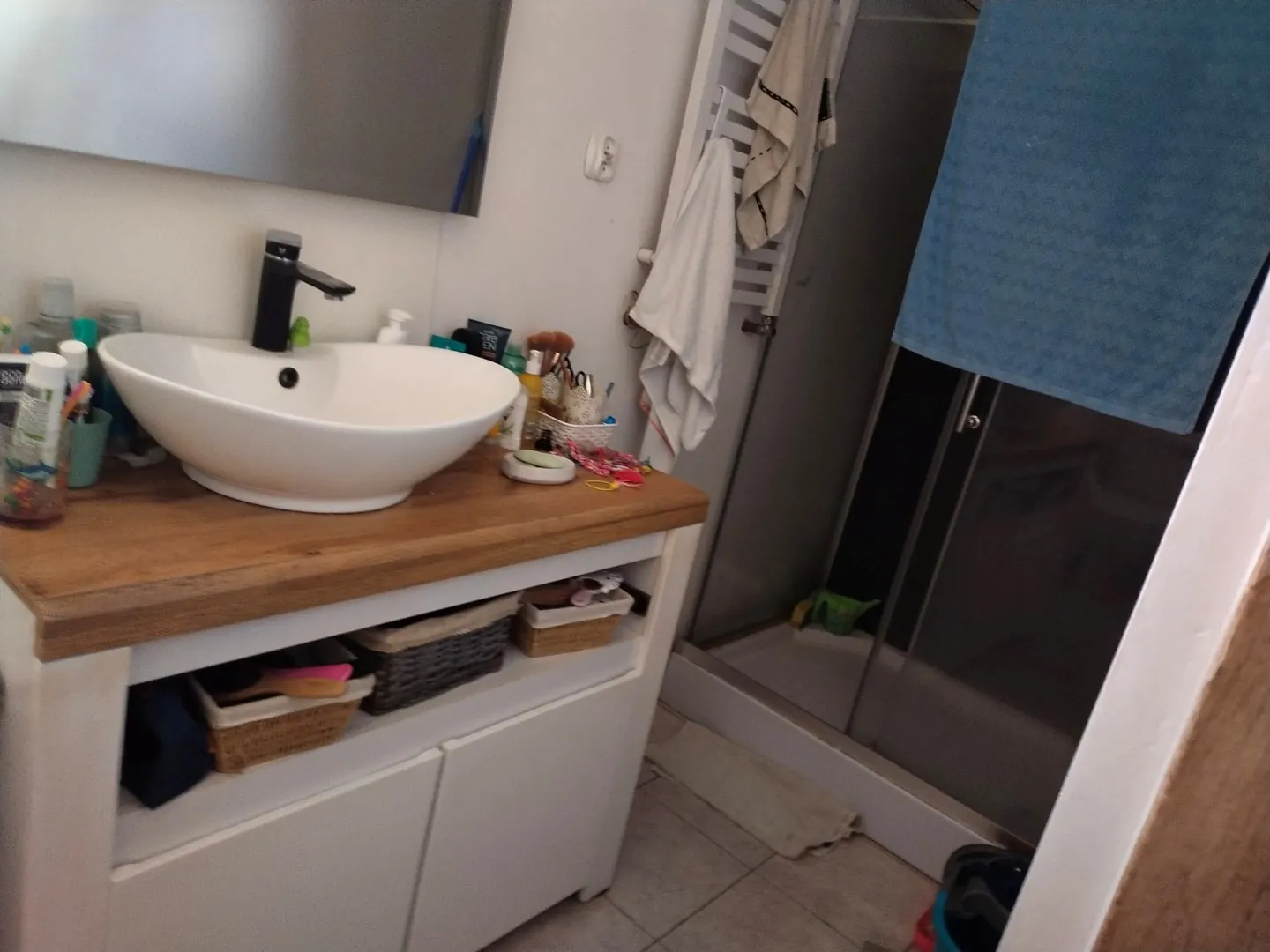 Wnętrze łazienki z białą, owalną umywalką nablatową ustawioną na drewnianym blacie. Pod blatem znajduje się biała szafka z otwartą półką, na której leżą wiklinowe koszyki i niebieska torba. Po prawej stronie widoczna jest kabina prysznicowa z przezroczystymi drzwiami i niebieską zasłoną w górnej części. Obok prysznica znajduje się biały grzejnik z powieszonym białym ręcznikiem. Na blacie obok umywalki stoją różnorodne kosmetyki i akcesoria toaletowe.