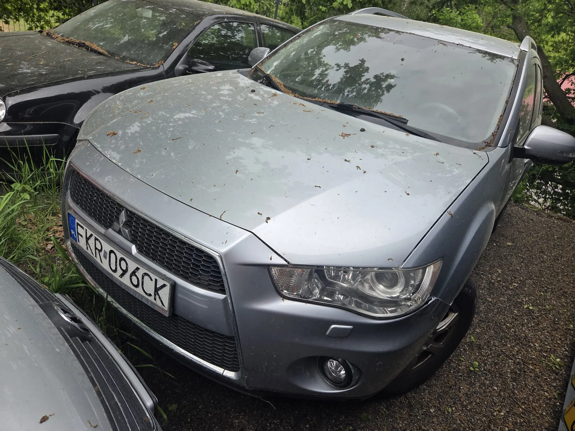 Samochód Mitsubishi w kolorze szarym z tablicą rejestracyjną FKR 096CK, zaparkowany na podłożu z żwiru. Na masce widoczne są plamy i resztki liści. W tle czarny samochód oraz zielone krzewy i drzewa. Po lewej stronie fragment innego pojazdu.