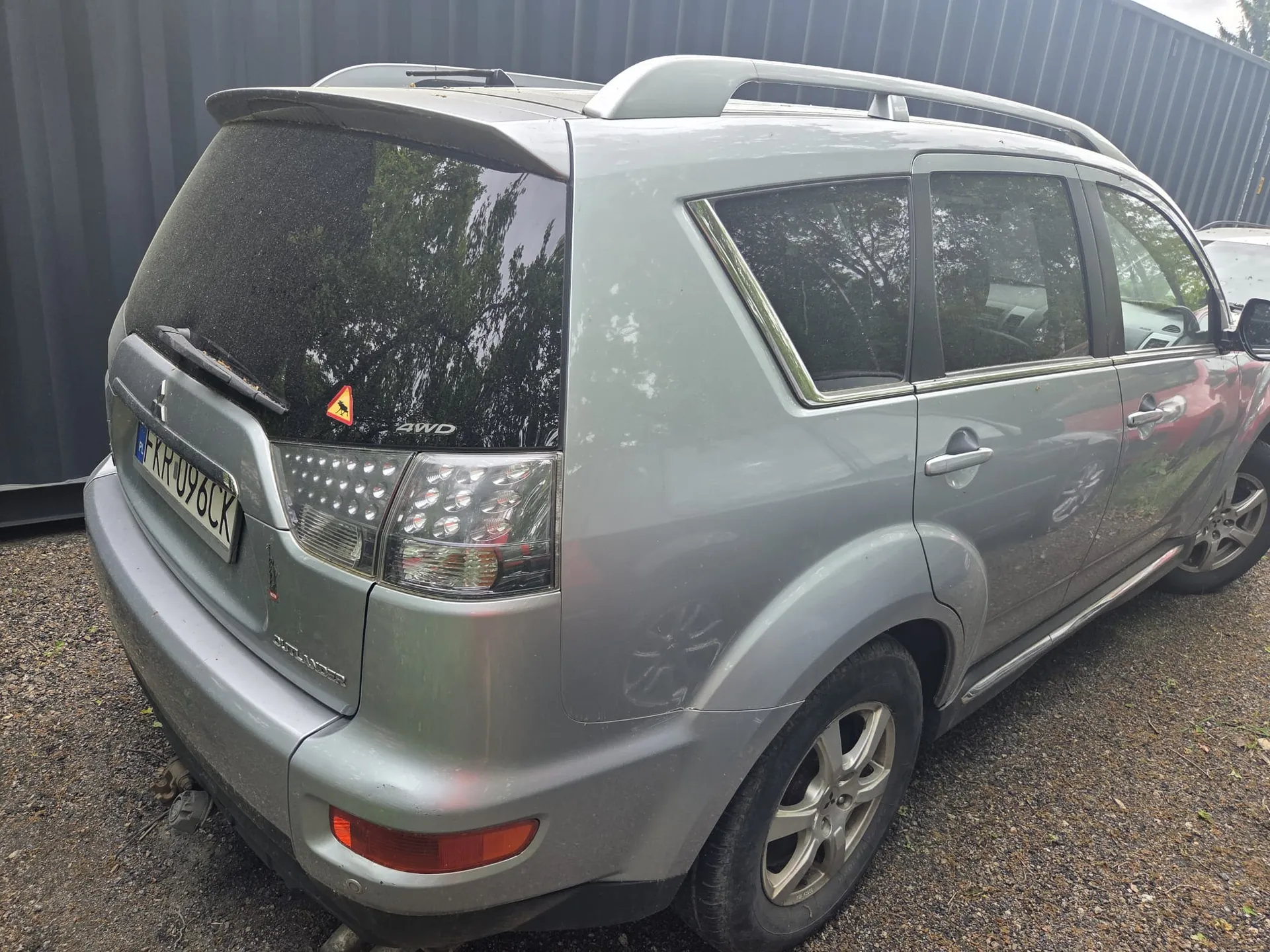 Srebrny SUV Mitsubishi Outlander z widocznym tyłem i bokiem, zaparkowany na żwirowym podłożu. Na tylnej szybie widoczny napis 4WD oraz trójkątny znak ostrzegawczy z czarnym symbolem zwierzęcia. Tablica rejestracyjna F-KR-096CK. Samochód wyposażony w dachowy bagażnik i chromowane listwy okienne. W tle ciemnoszara metalowa ogrodzenie i fragment innego pojazdu po prawej stronie.