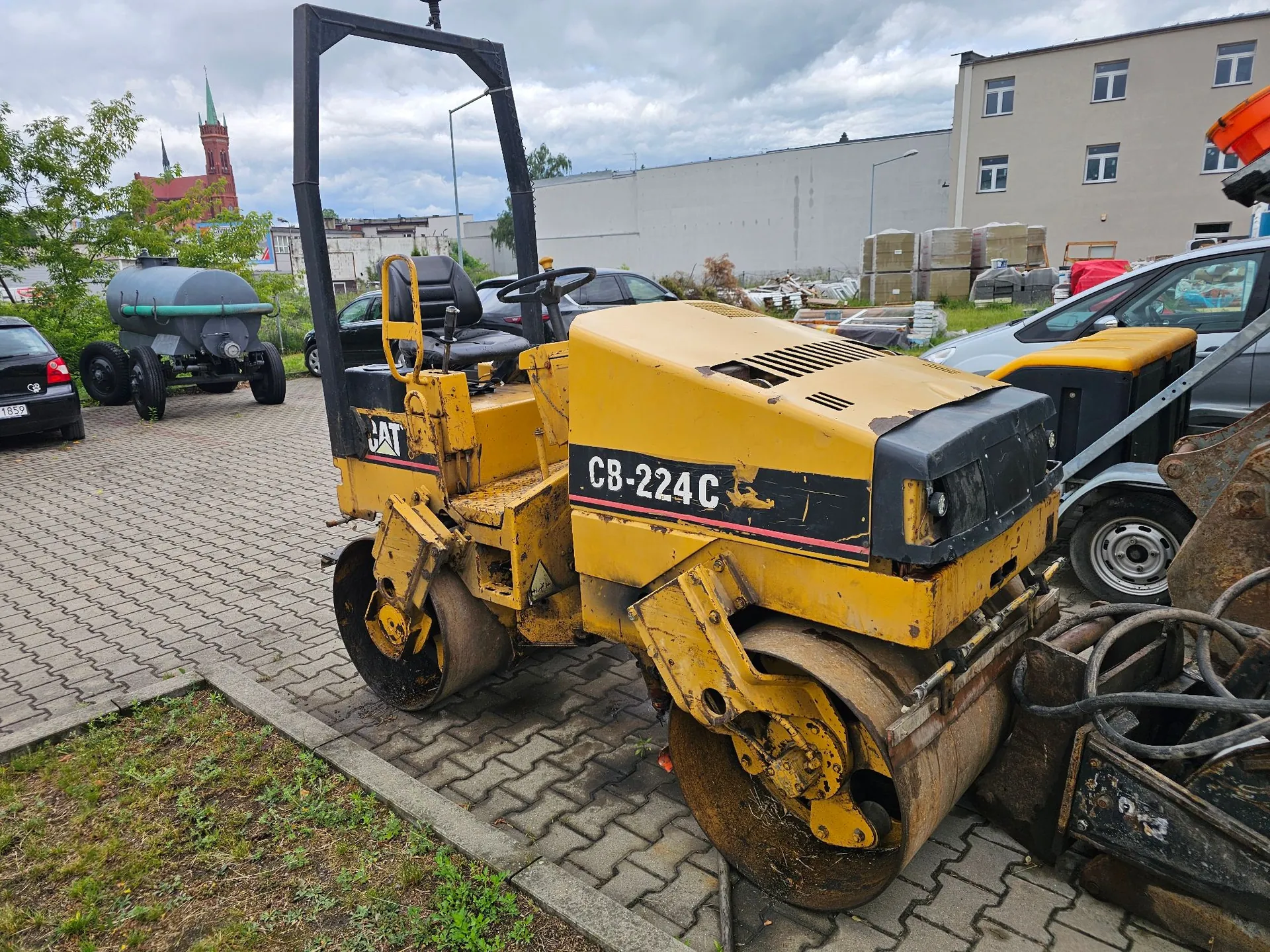 Żółty walec drogowy marki CAT, model CB-224C, zaparkowany na kostce brukowej. Maszyna posiada dwa metalowe bębny i otwartą kabinę z czarnym fotelem i stelażem ochronnym. W tle widoczny jest szary budynek, ciemny samochód osobowy oraz zielony zbiornik na kołach. W oddali widać wieżę kościoła.