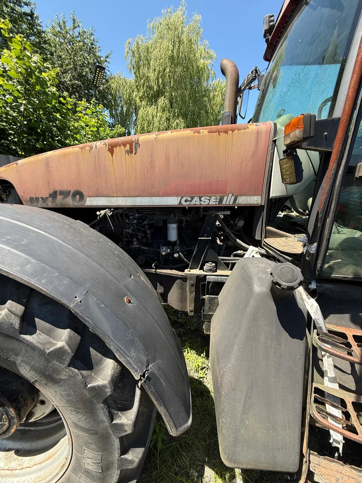 Zbliżenie na przednią część starego, zardzewiałego traktora marki CASE IH. Widoczna jest maska z widoczną korozją, duże przednie koło z czarnym błotnikiem oraz fragment kabiny z lusterkiem i kierunkowskazem. W tle znajdują się zielone drzewa i niebieskie niebo.